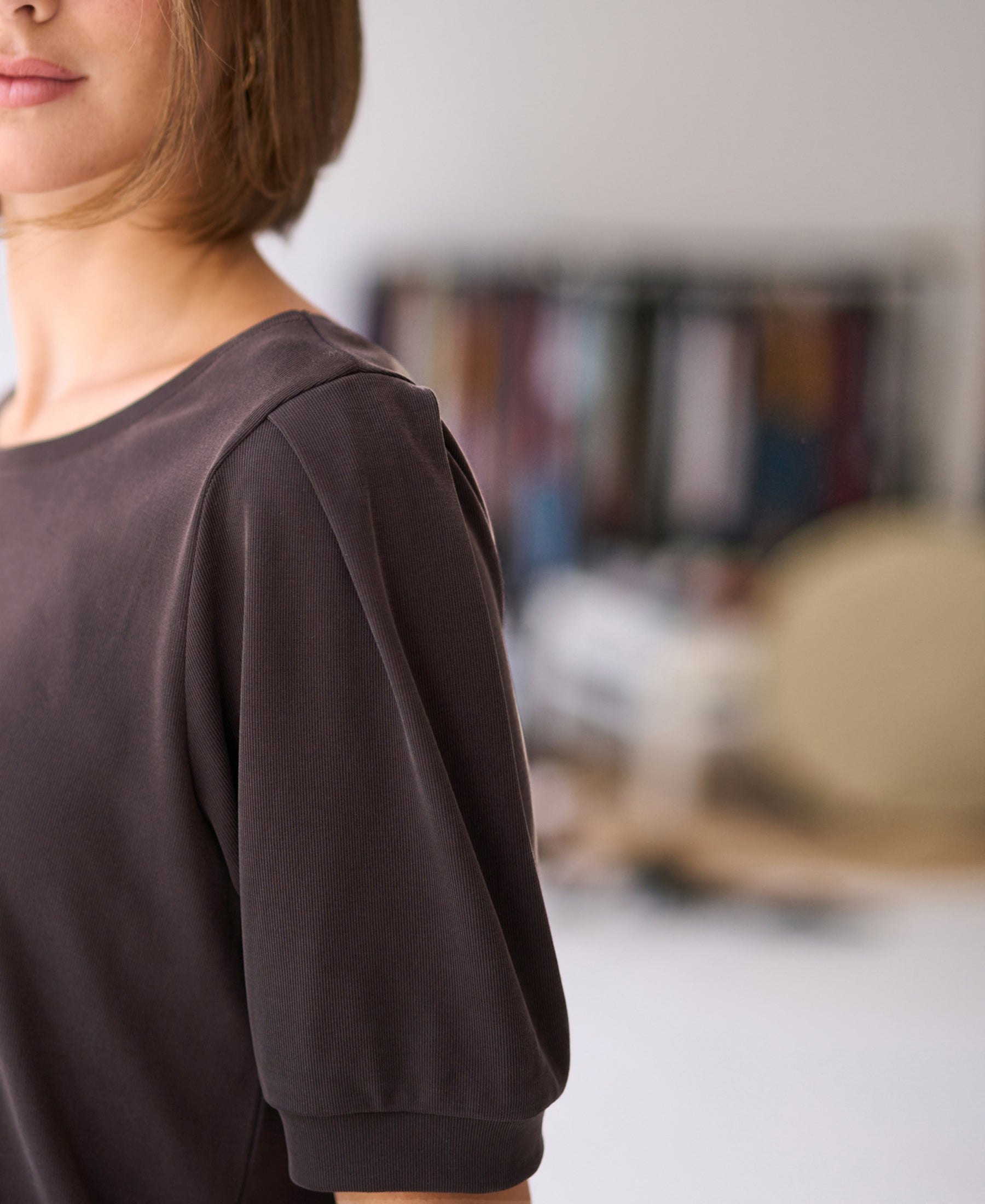Woman wearing a ribbed chocolate brown T-shirt indoors, with colorful fabrics softly blurred in the background.