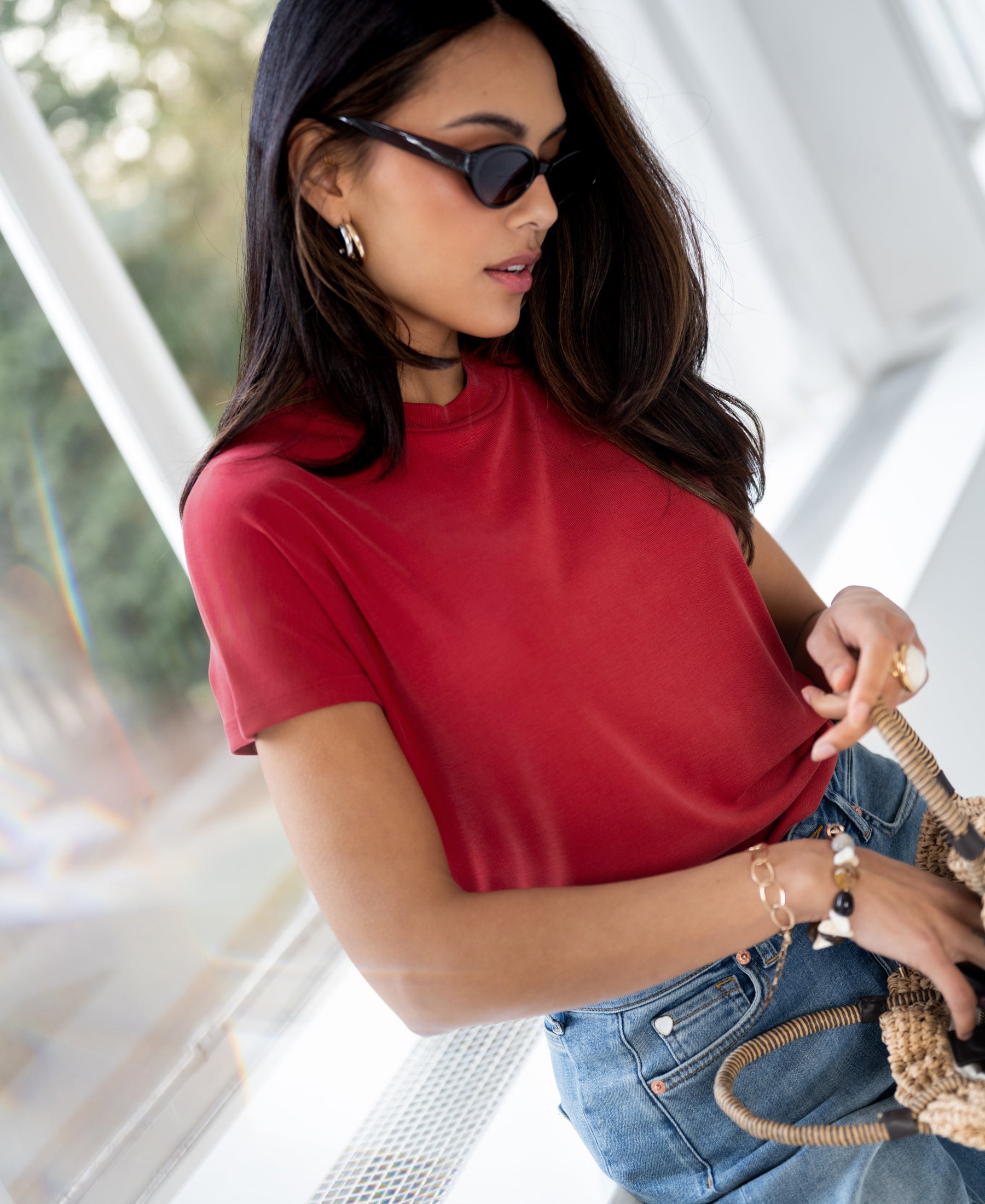 Woman wearing a red ribbed top looks into a woven bag by a window.