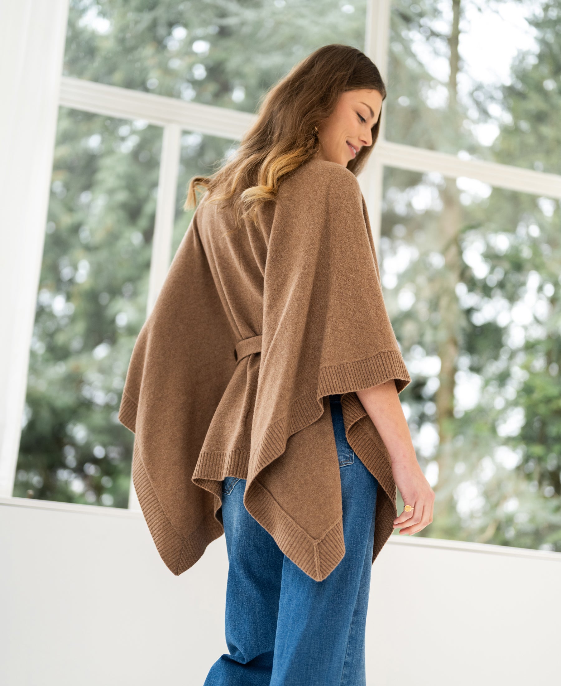 Camel merino wool poncho by Plein Publique, shown on a woman standing by a window and smiling softly.