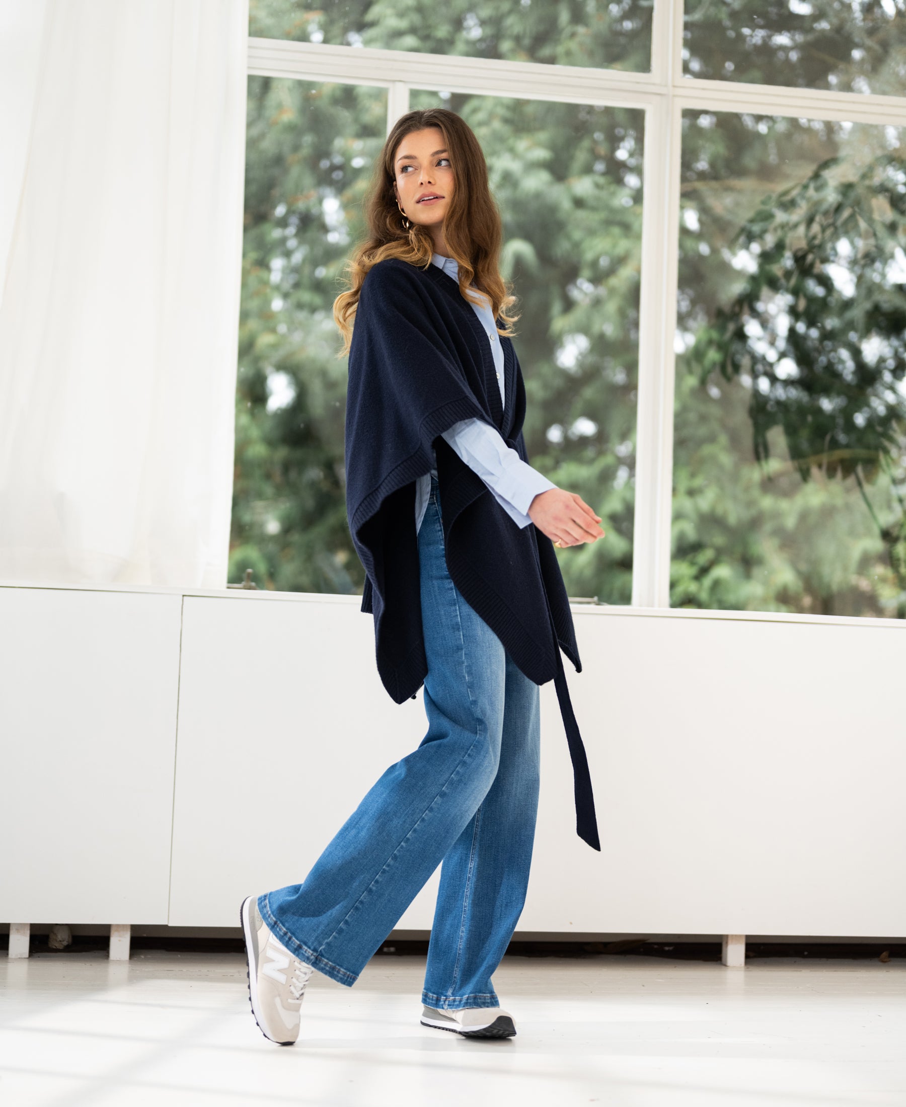 A dark blue merino wool poncho by Plein Publique displayed near sunlit windows with greenery in the background.