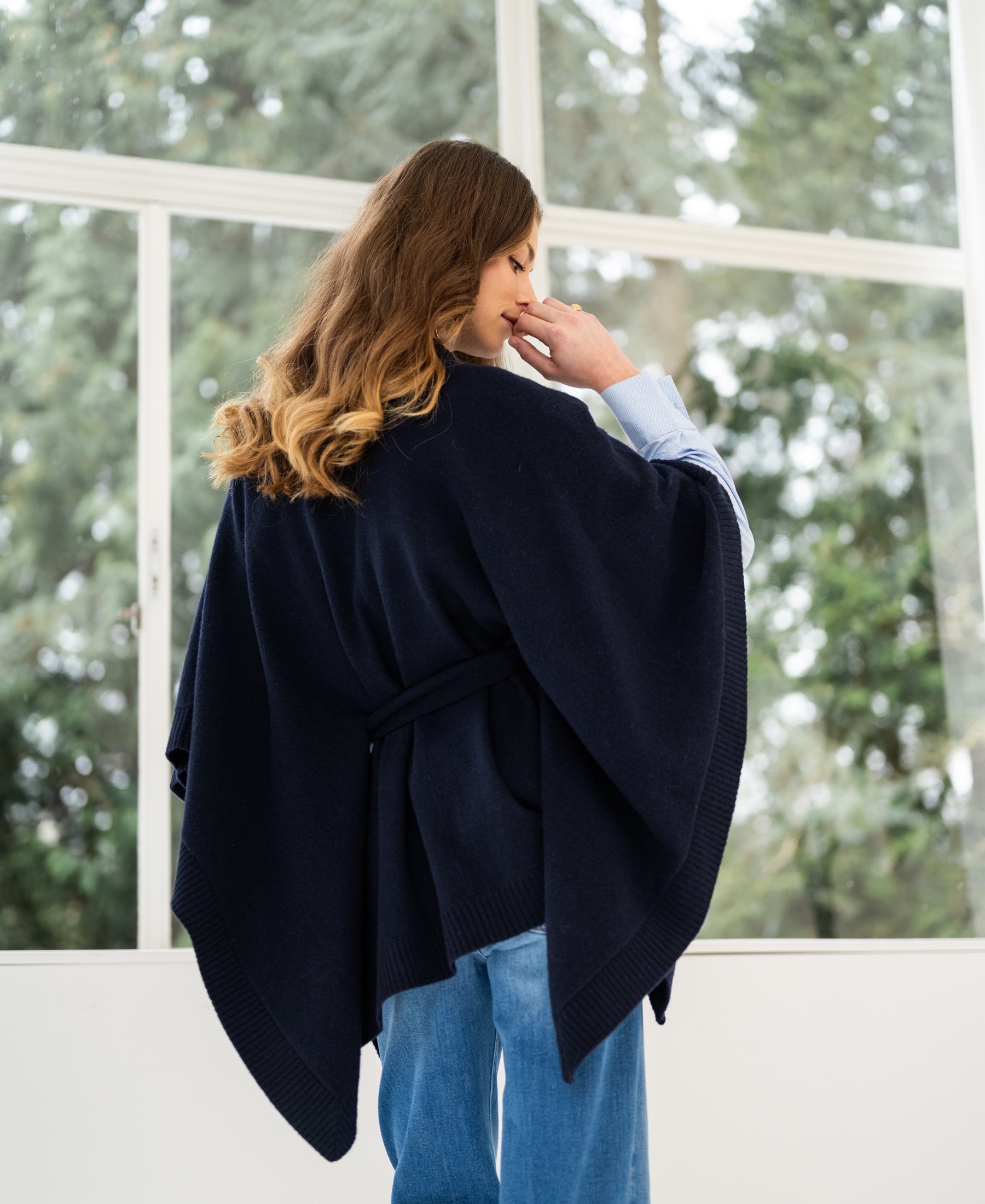 Navy blue merino wool poncho by PLEIN PUBLIQUE, shown from the back near large windows.