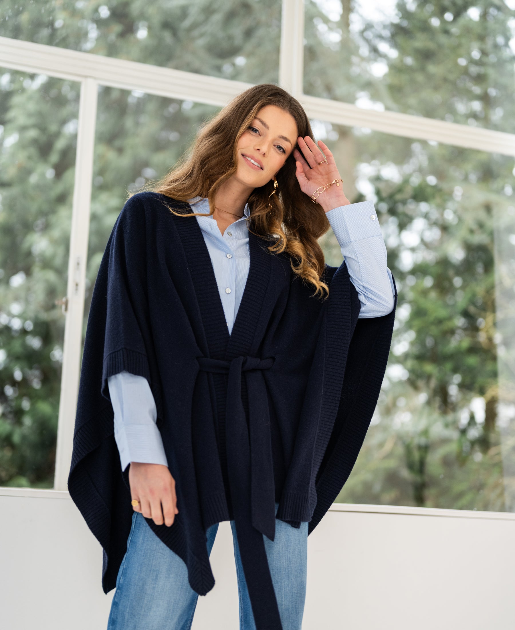 Woman wears the PLEIN PUBLIQUE merino wool poncho in dark blue indoors by large windows.