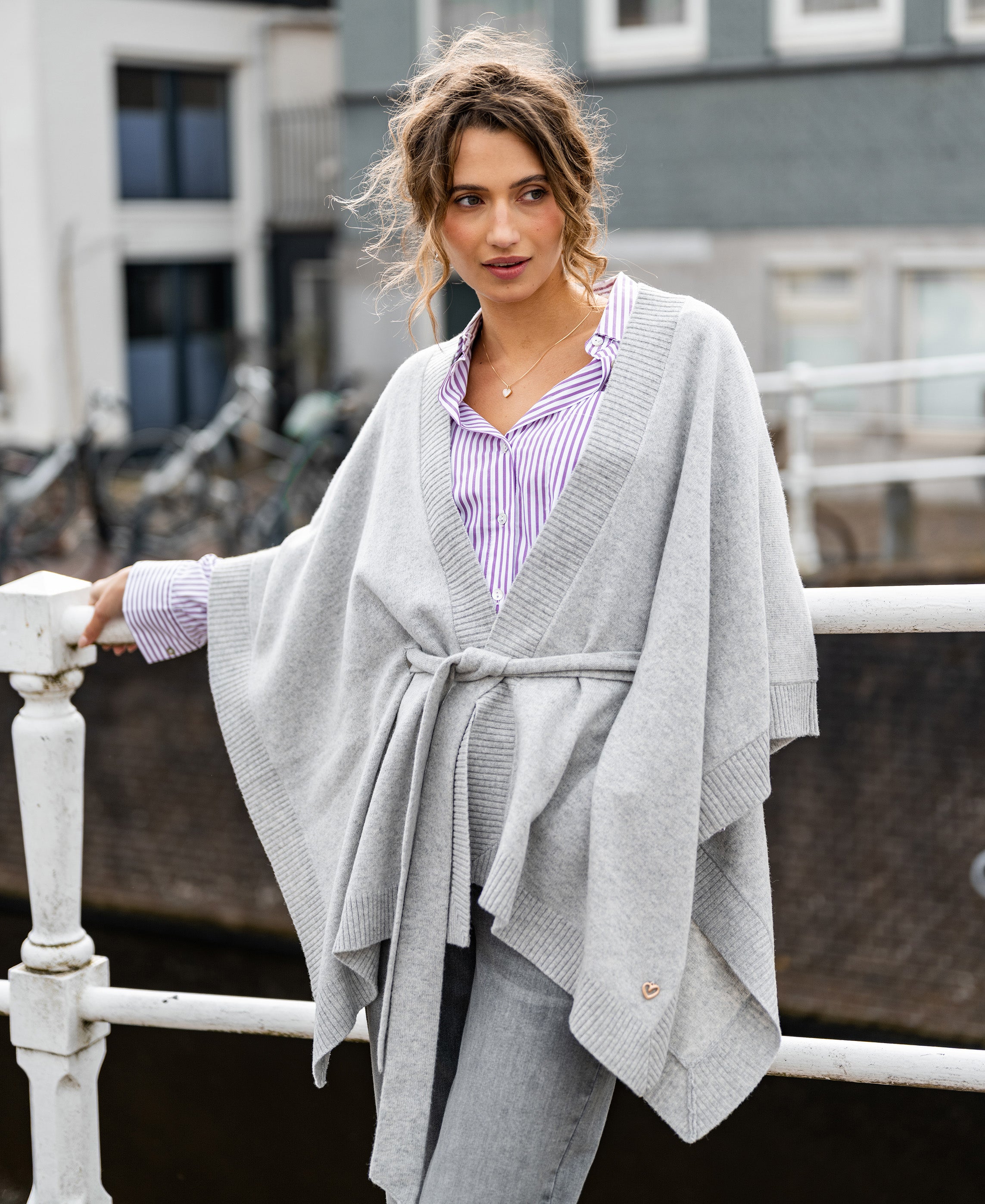 Woman wearing a silver-grey merino wool poncho stands on a bridge, with bikes visible in the background.