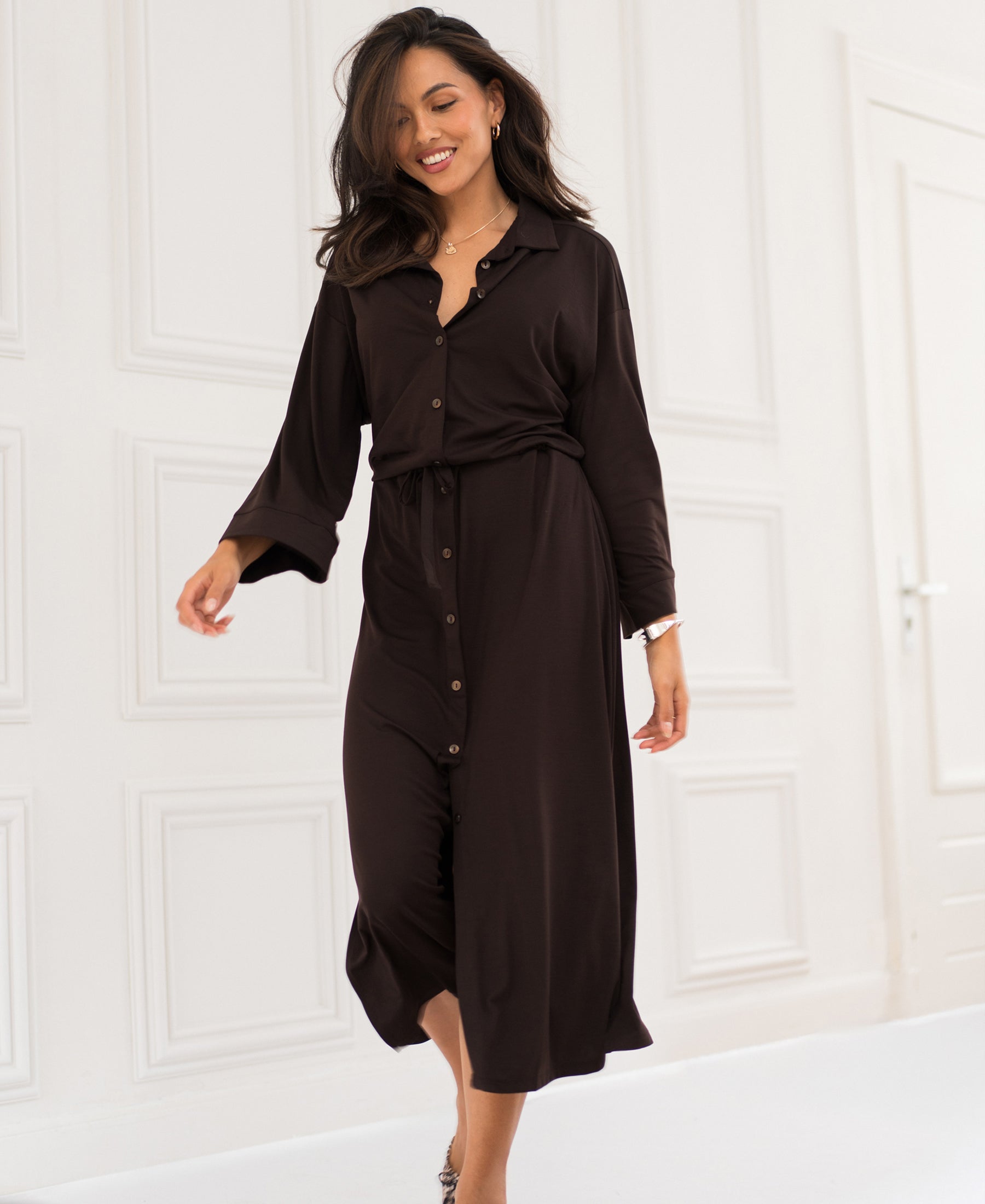 Front button dress LE RAPHAEL in chocolate brown by PLEIN PUBLIQUE, shown on a model in a white minimalistic room.