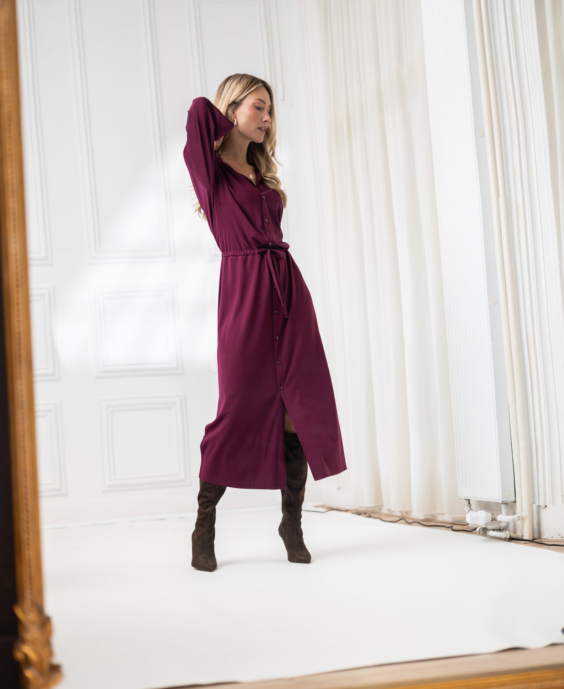 Woman wears Bordeaux LE RAPHAEL button-down dress by PLEIN PUBLIQUE in a bright, elegant room.