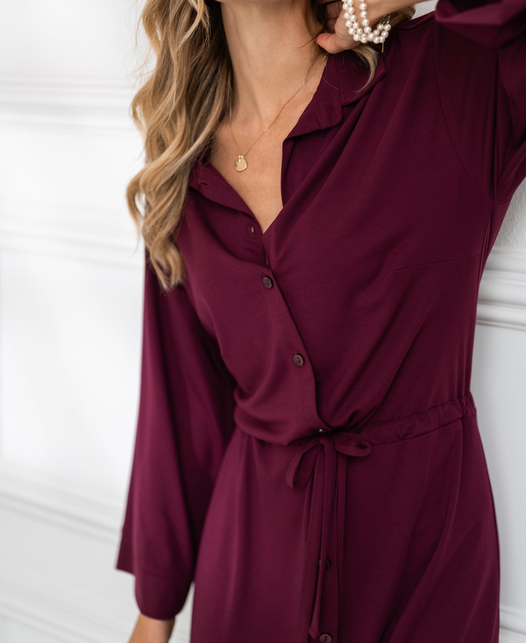 Front view of a woman wearing a Bordeaux button-down dress by PLEIN PUBLIQUE, standing against a white wall.
