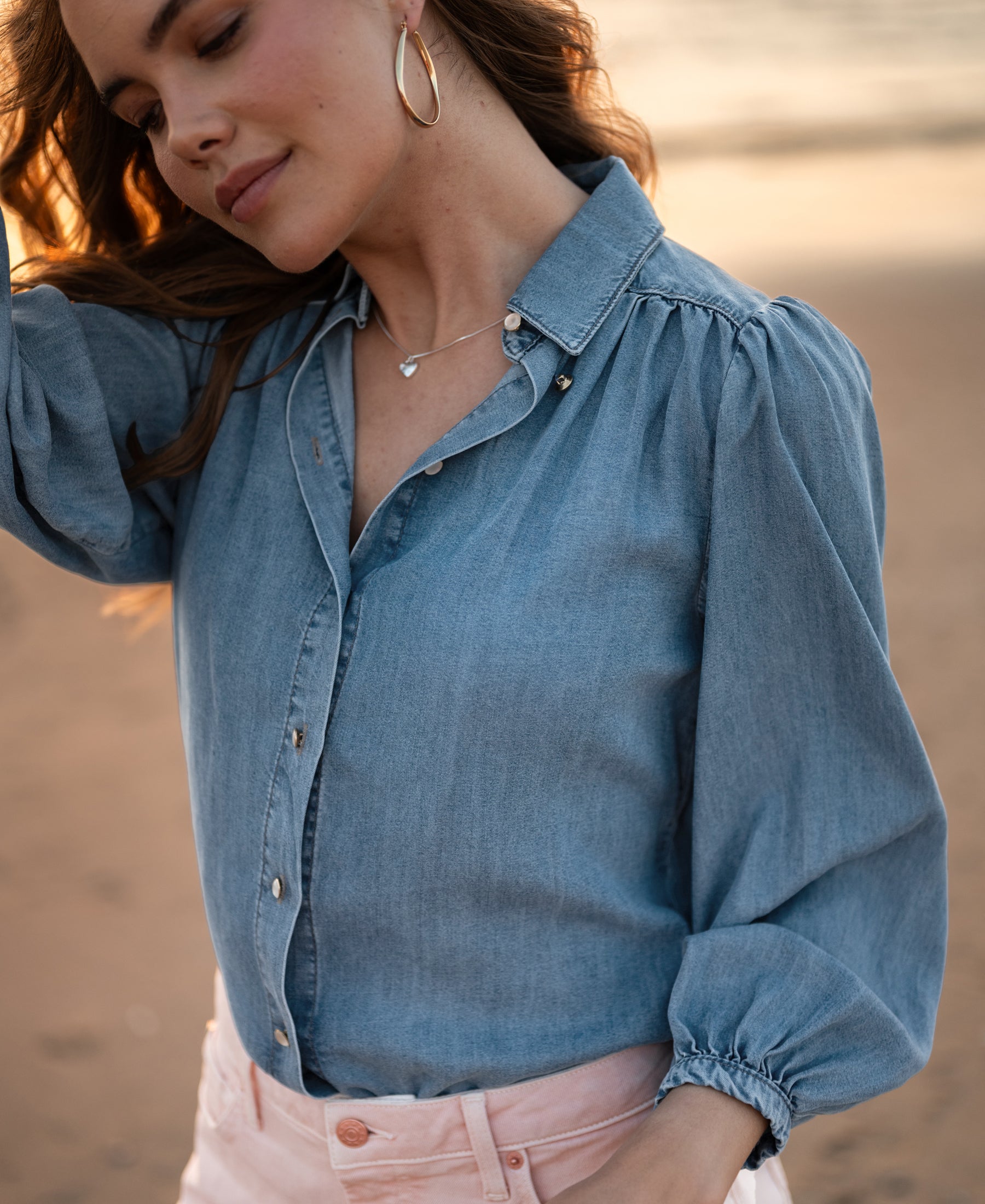 Woman wearing LA RIVA Stonewash Tencel denim blouse by PLEIN PUBLIQUE, wind blowing her hair on the beach.