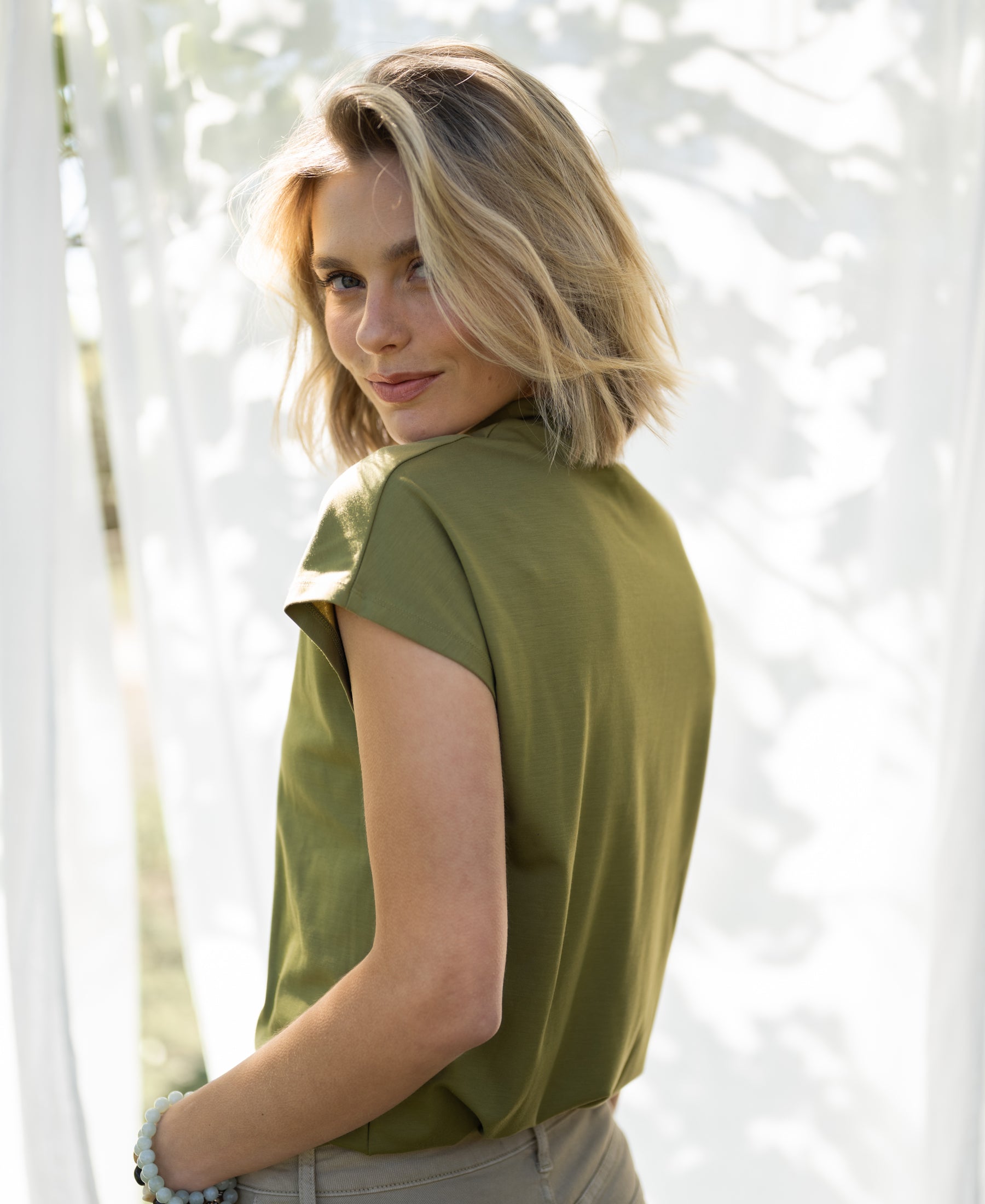 Olive green LA ROMY travel collared top by PLEIN PUBLIQUE shown in sunlight near white curtains.