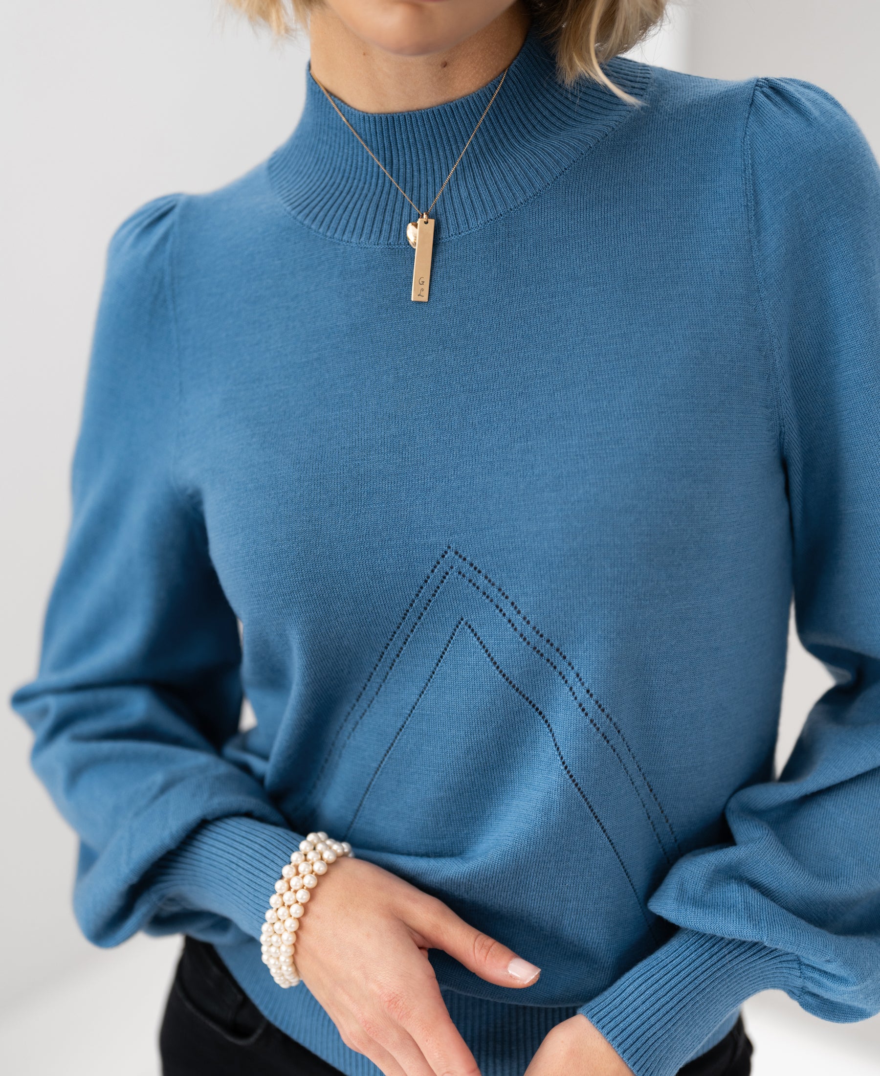 Front view of a woman wearing a steel blue PLEIN PUBLIQUE LES SALLES turtleneck sweater, hands clasped.