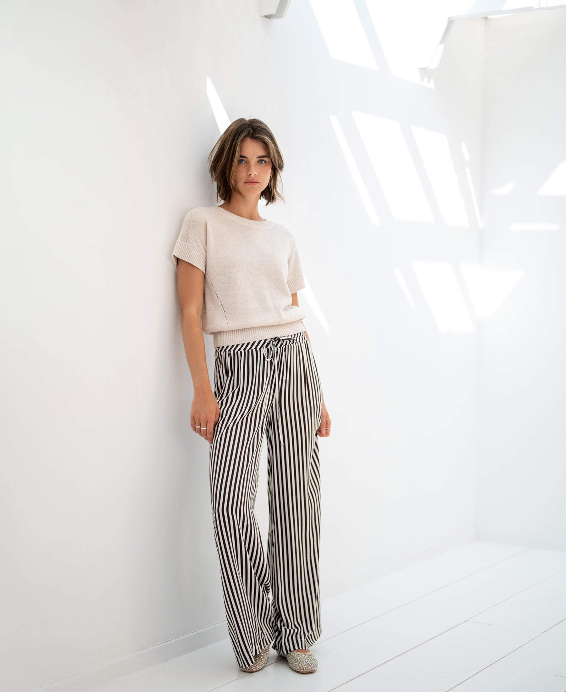 Woman wearing PLEIN PUBLIQUE linen summer sweater LE SOLEIL in light sand, shown in a bright white room.