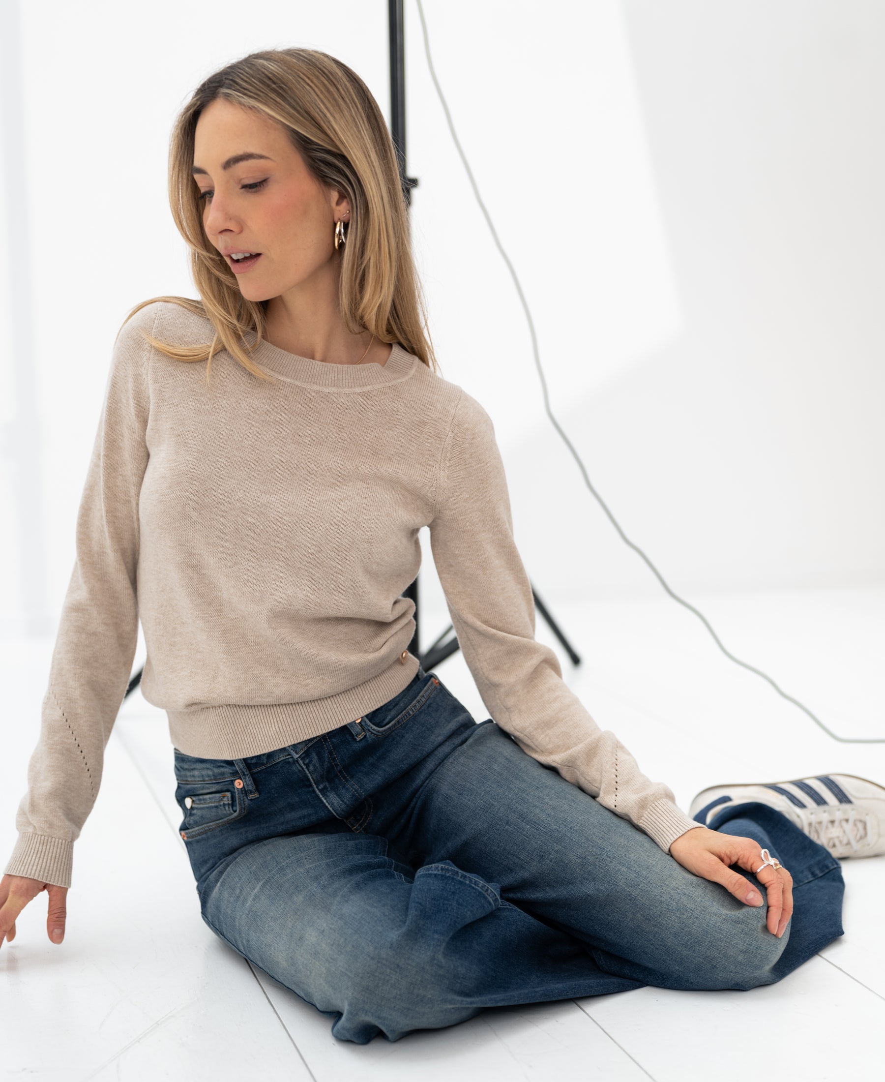Woman wearing the Merino sweater LE SONGE Sand from PLEIN PUBLIQUE, seated on the floor in a bright studio.
