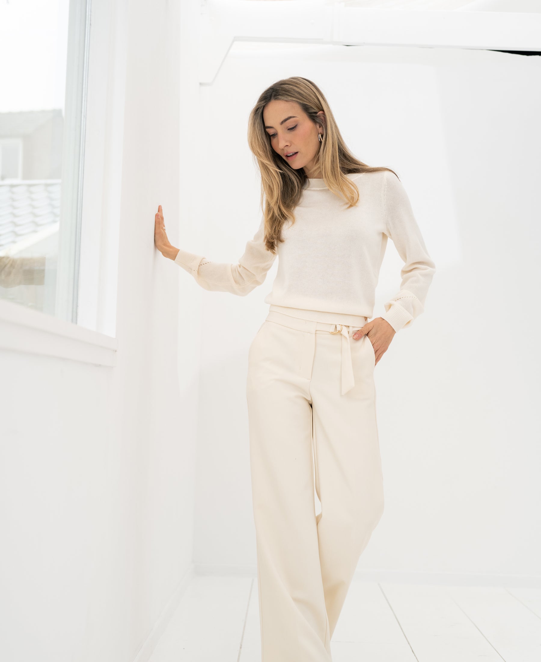 Cream LA WENDE pants by PLEIN PUBLIQUE, shown on a woman standing by a window, viewed from the front.