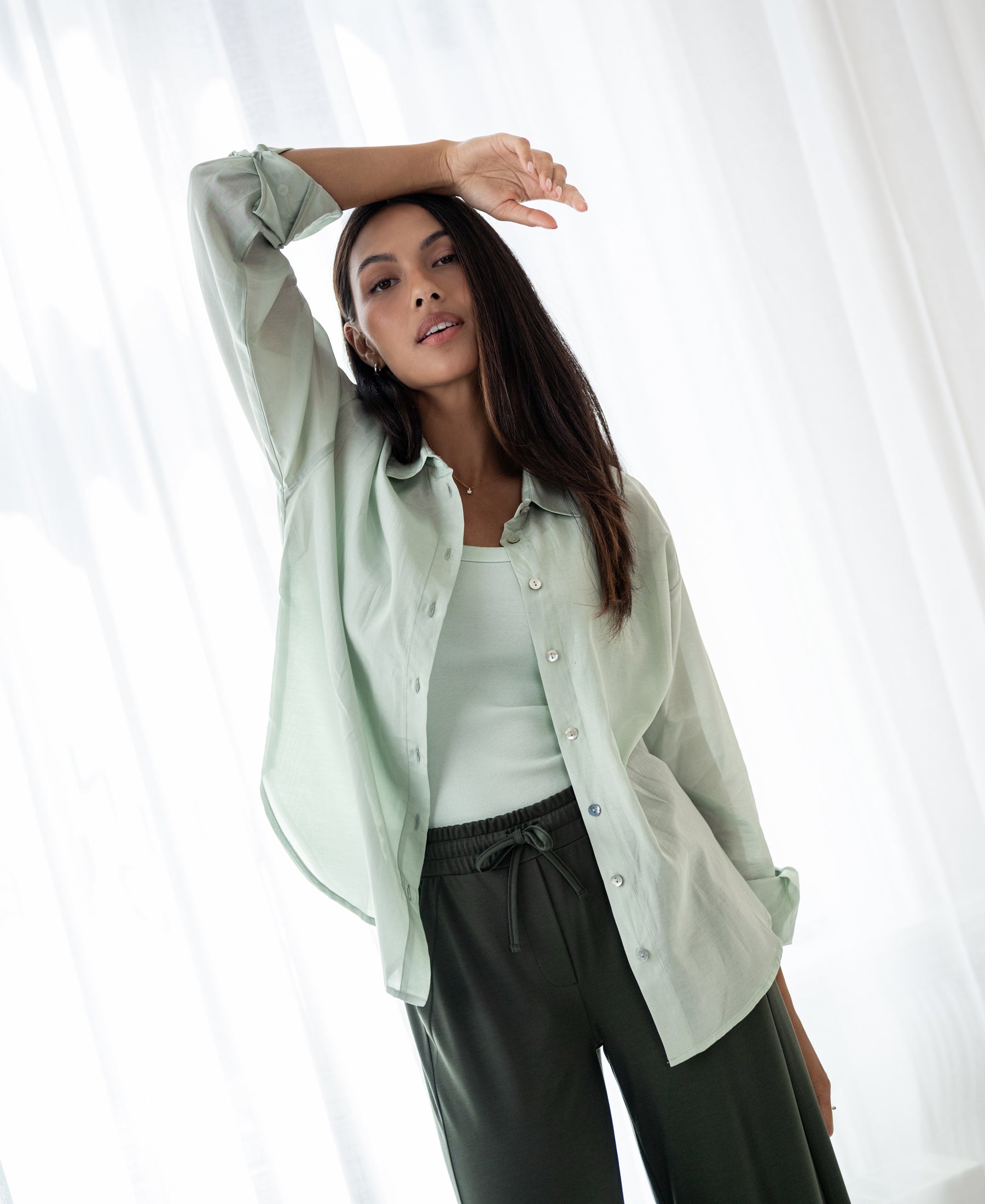 Woman models the PLEIN PUBLIQUE Dunne oversized light green silk blouse by sheer white curtains indoors.