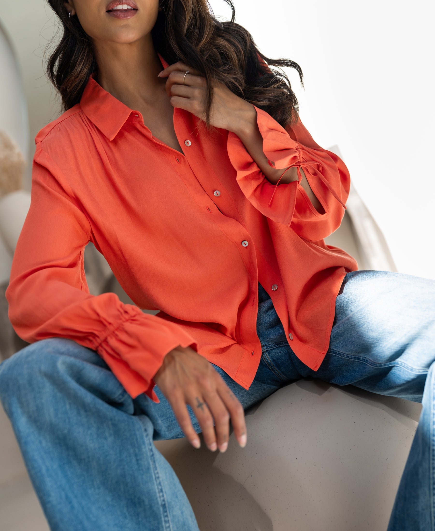 Coral LA TROYES blouse by PLEIN PUBLIQUE, shown worn with one hand on the collar and the other resting on her leg.