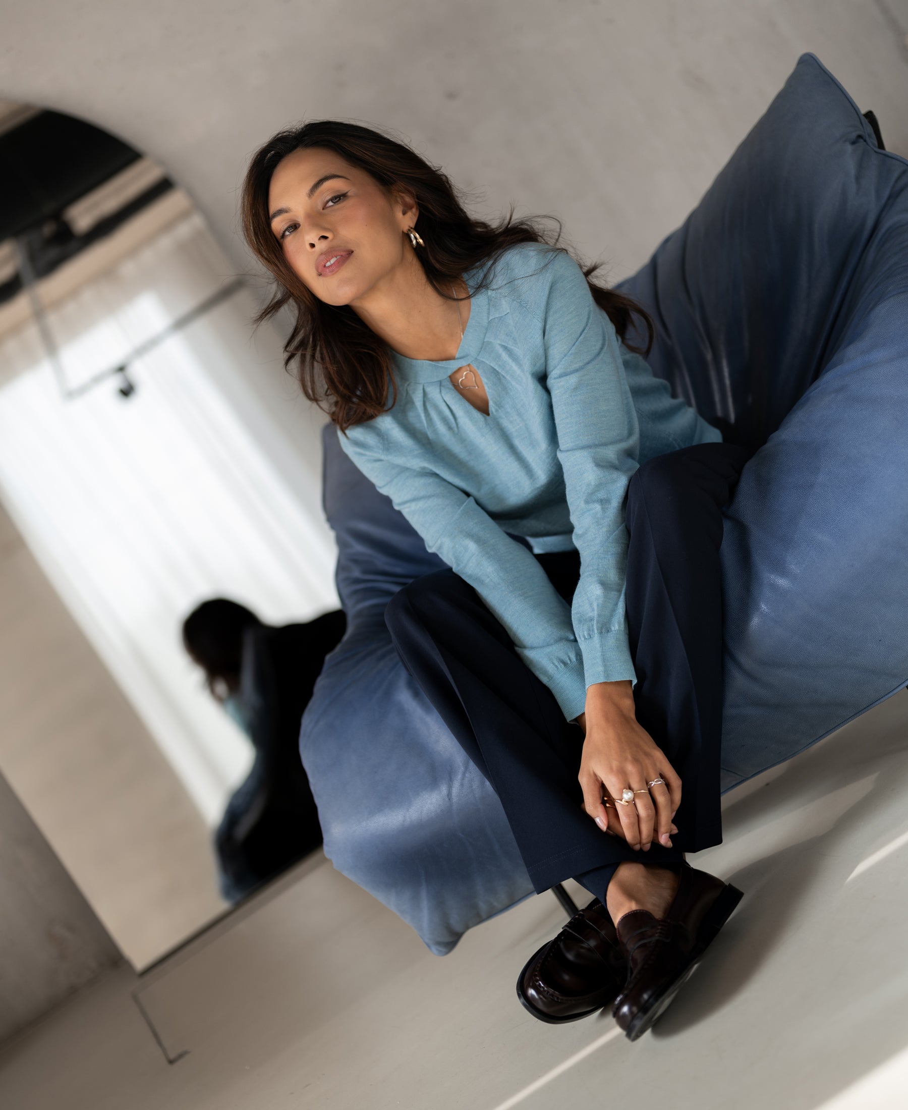 Woman wearing PLEIN PUBLIQUE Merino wool sweater LA TWIST in mist blue, seated; mirror reflection visible.