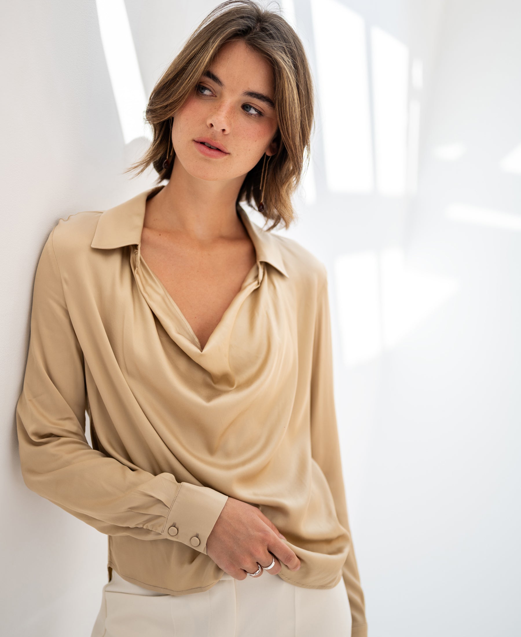 LE VENT satin stretch blouse in light sand by PLEIN PUBLIQUE, shown against a white wall in natural light.