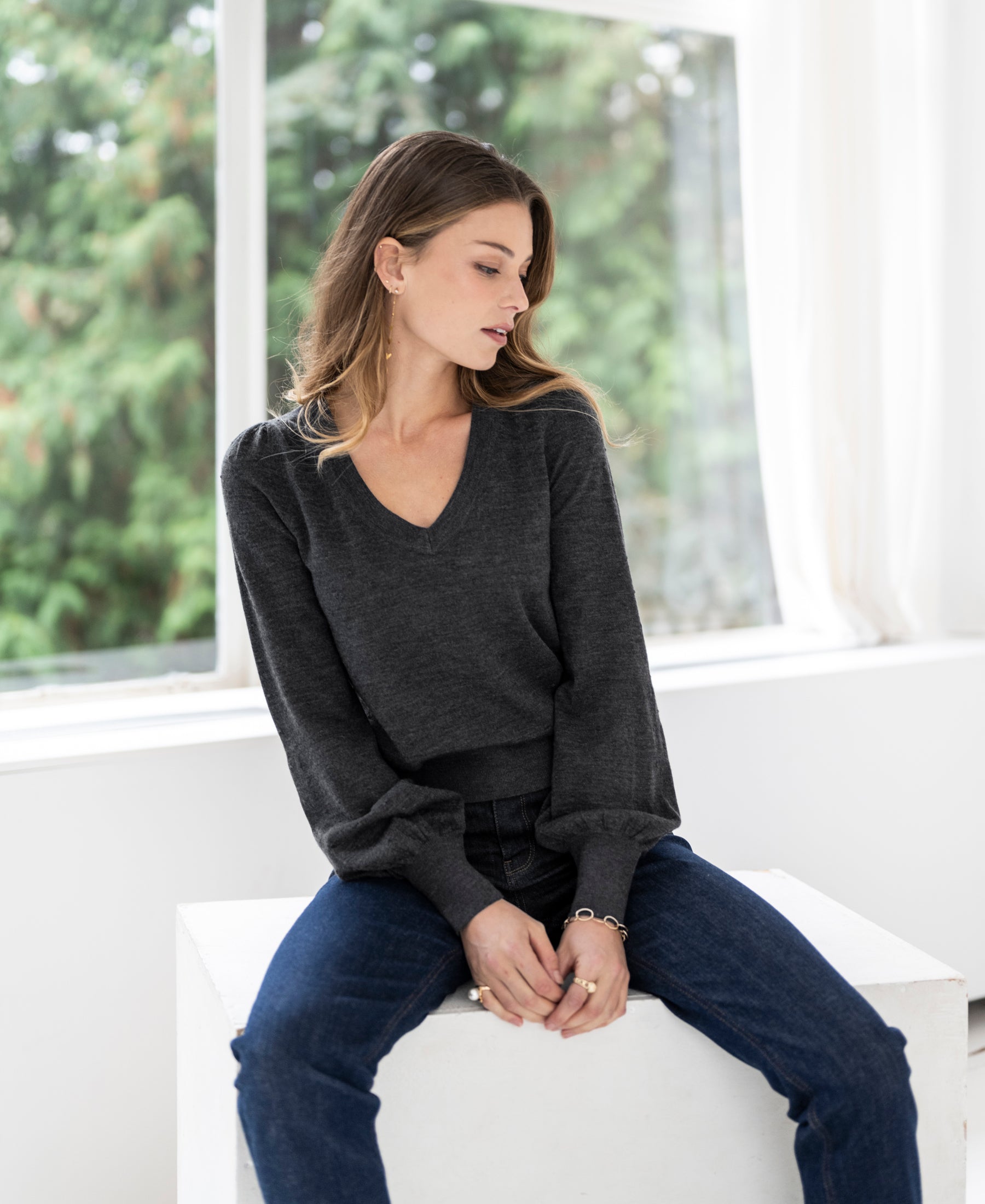 Woman wearing the Merino V-neck sweater LA VICTORIA Anthracite by PLEIN PUBLIQUE, sitting on a couch.