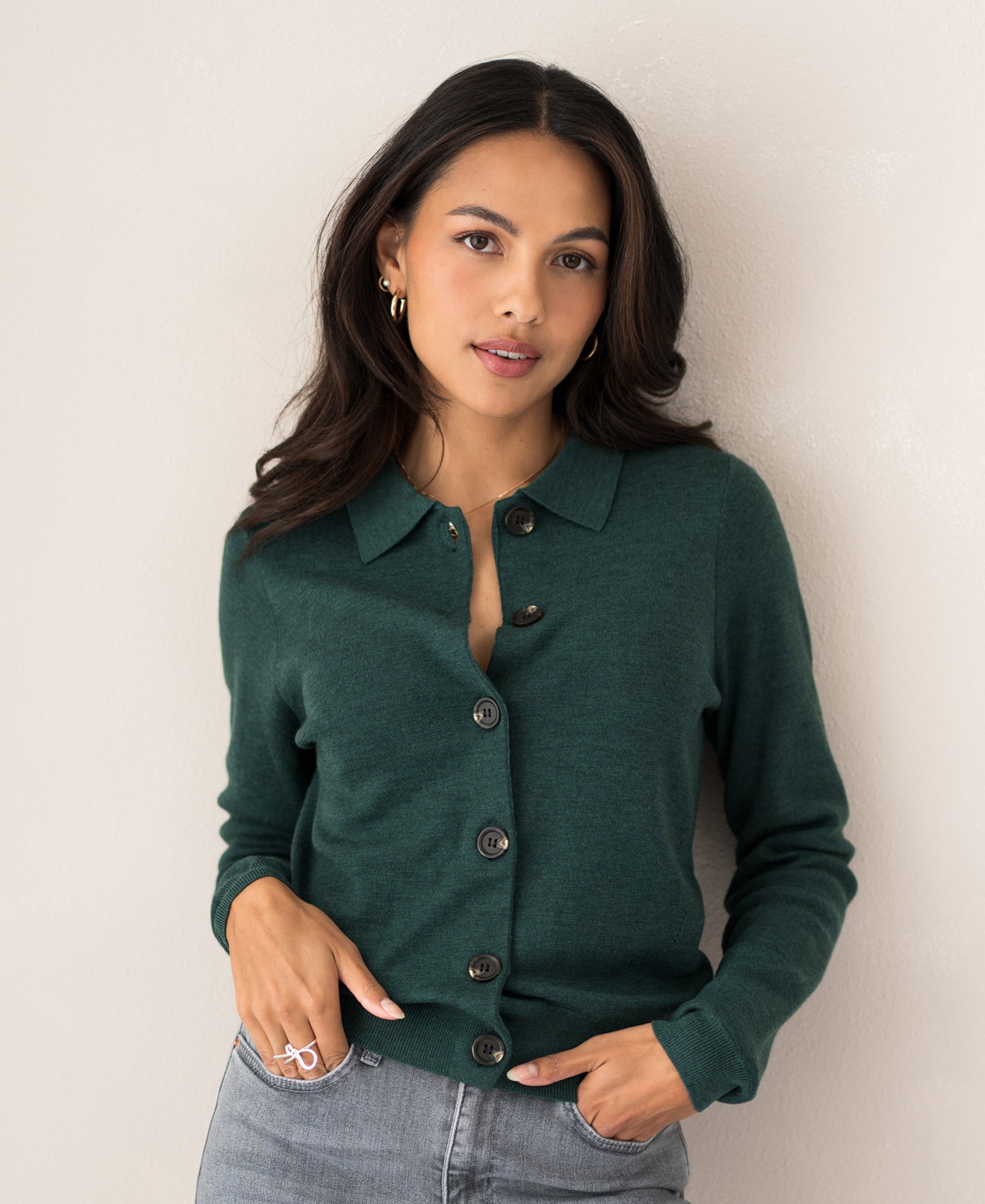 Woman wearing a greenish collared vest, standing by a light wall.