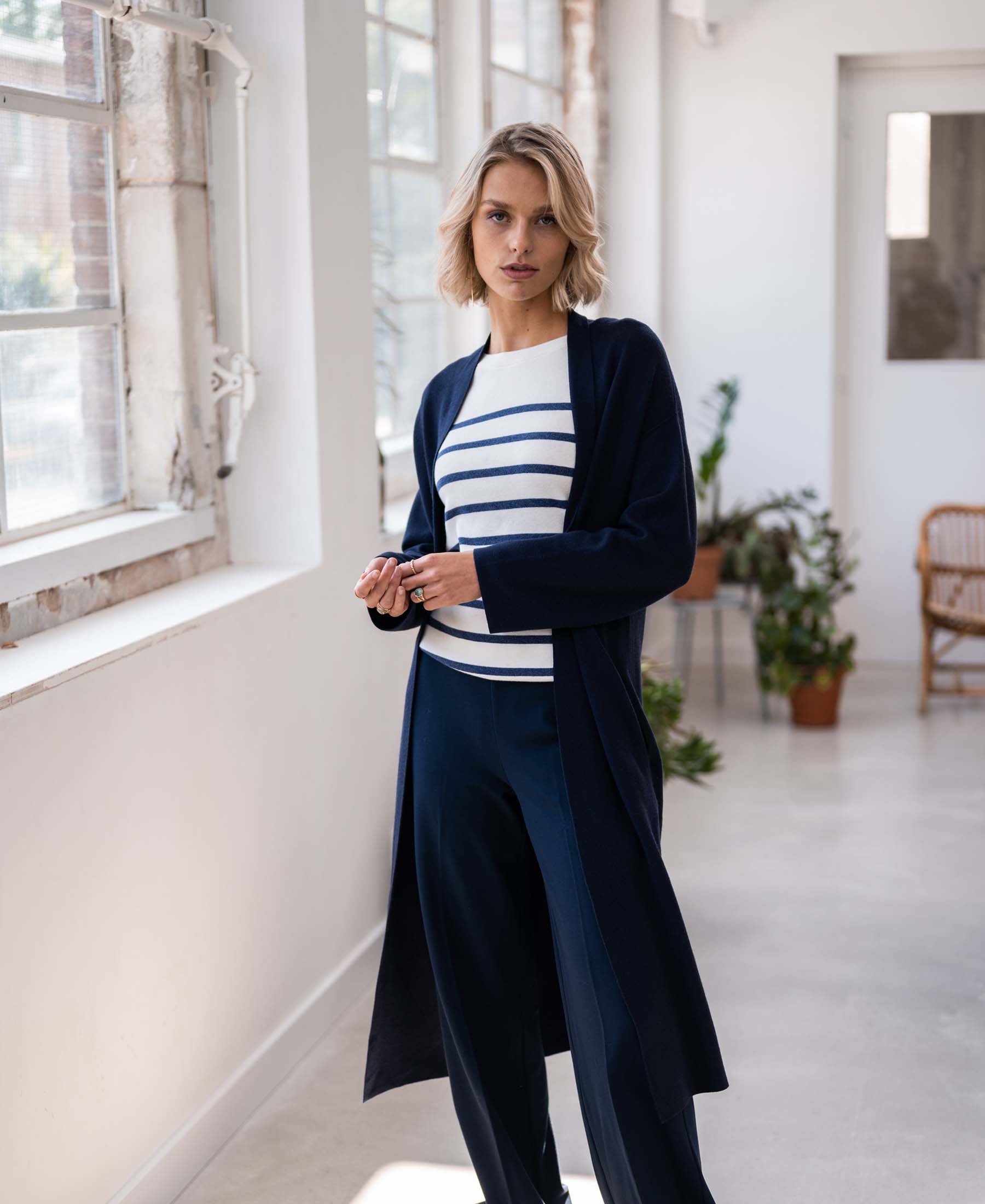 Woman wearing a PLEIN PUBLIQUE Breton sweater LELISA in ivory and denim blue stands by plants and large windows.