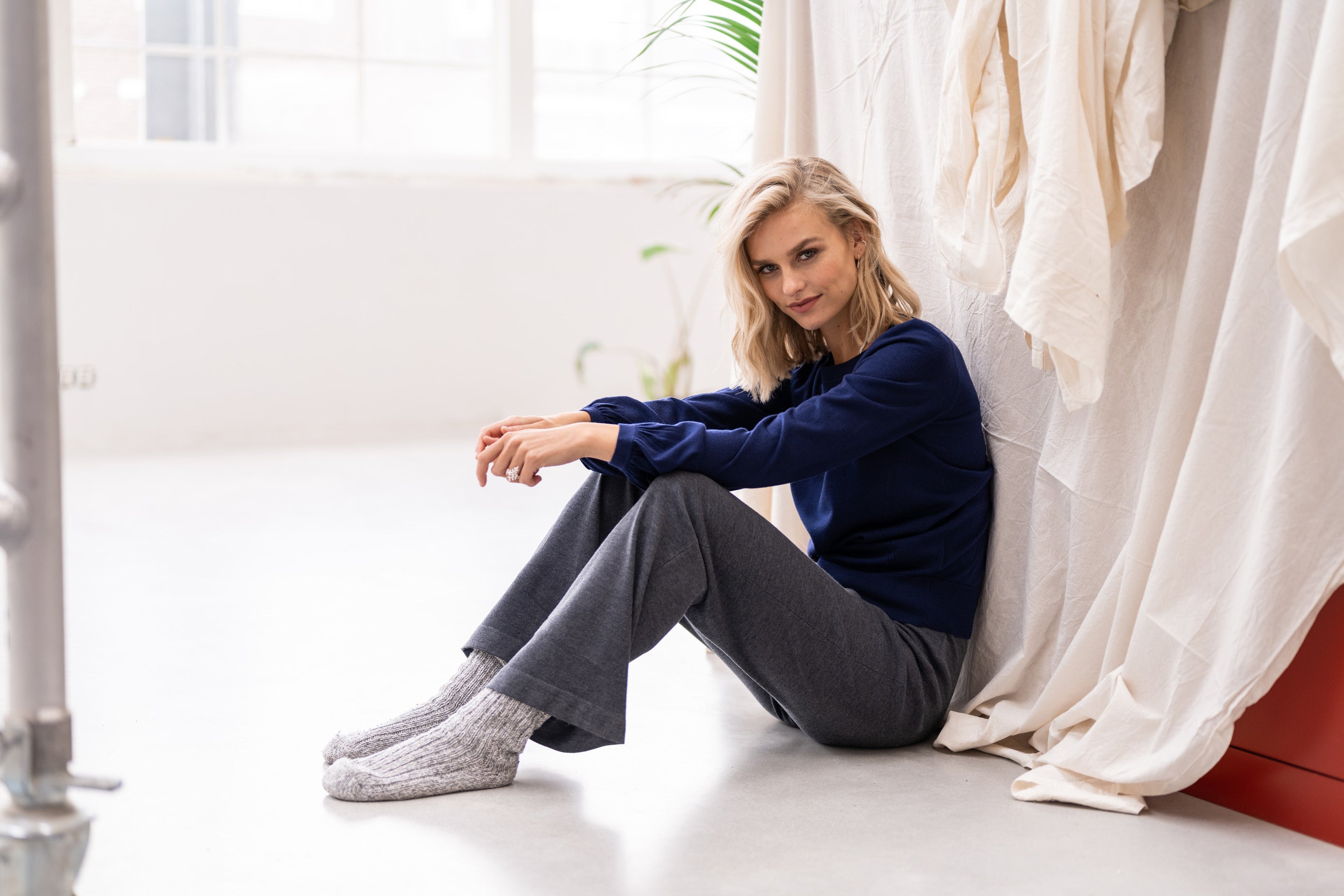 Grey knitted pants by PLEIN PUBLIQUE, LE LOURDES style, shown on a seated model in a bright room.