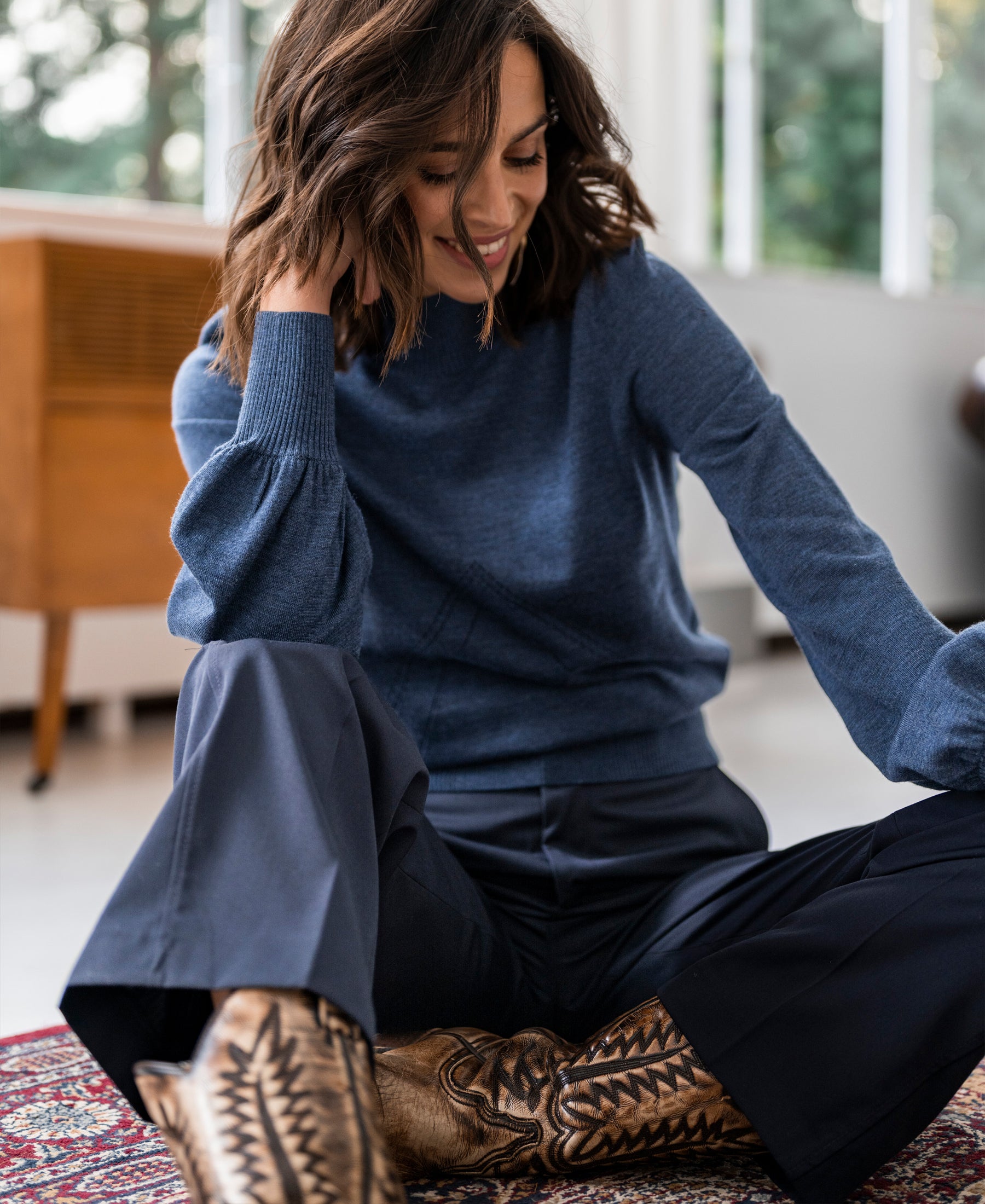 Woman indoors, smiling, wearing the PLEIN PUBLIQUE LES SALLES blue turtleneck sweater.