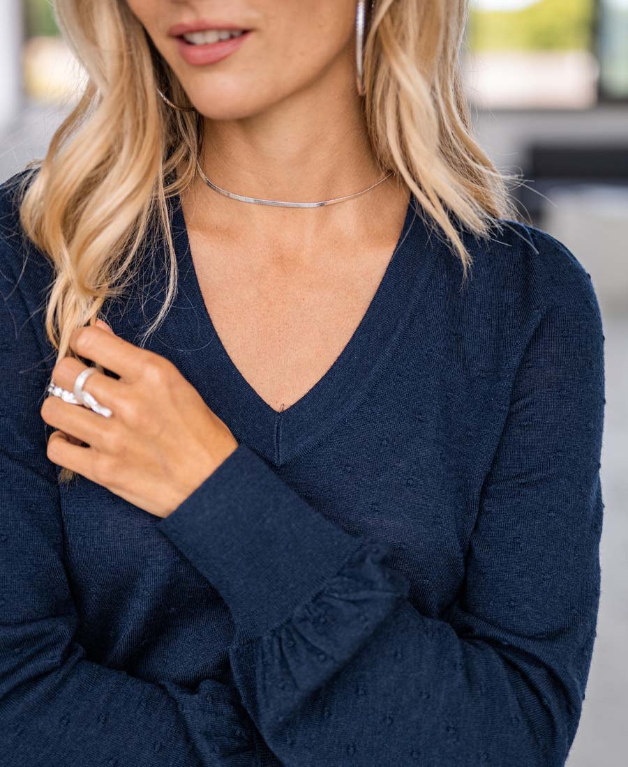 Woman with long blonde hair wearing the Merino V-hals sweater LA VICTORIA Donkerblauw, styled with a silver necklace and rings. Her face is partially visible, hand in her hair, against a blurred background—ideal for elegante dames knitwear fans.
