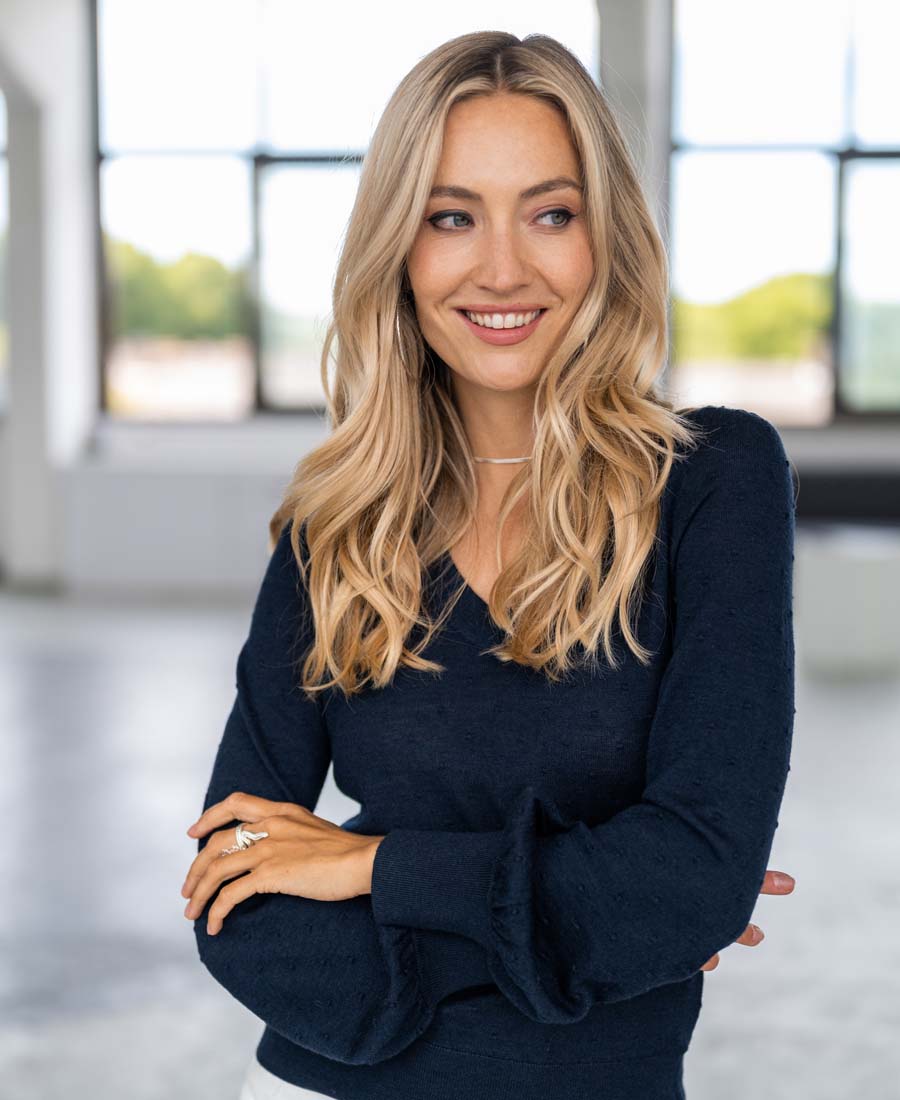 A woman with long blonde hair stands indoors, arms crossed and smiling to the side, wearing a Merino V-hals trui LA VICTORIA Donkerblauw. Large windows behind her reveal a blurred outdoor scene.