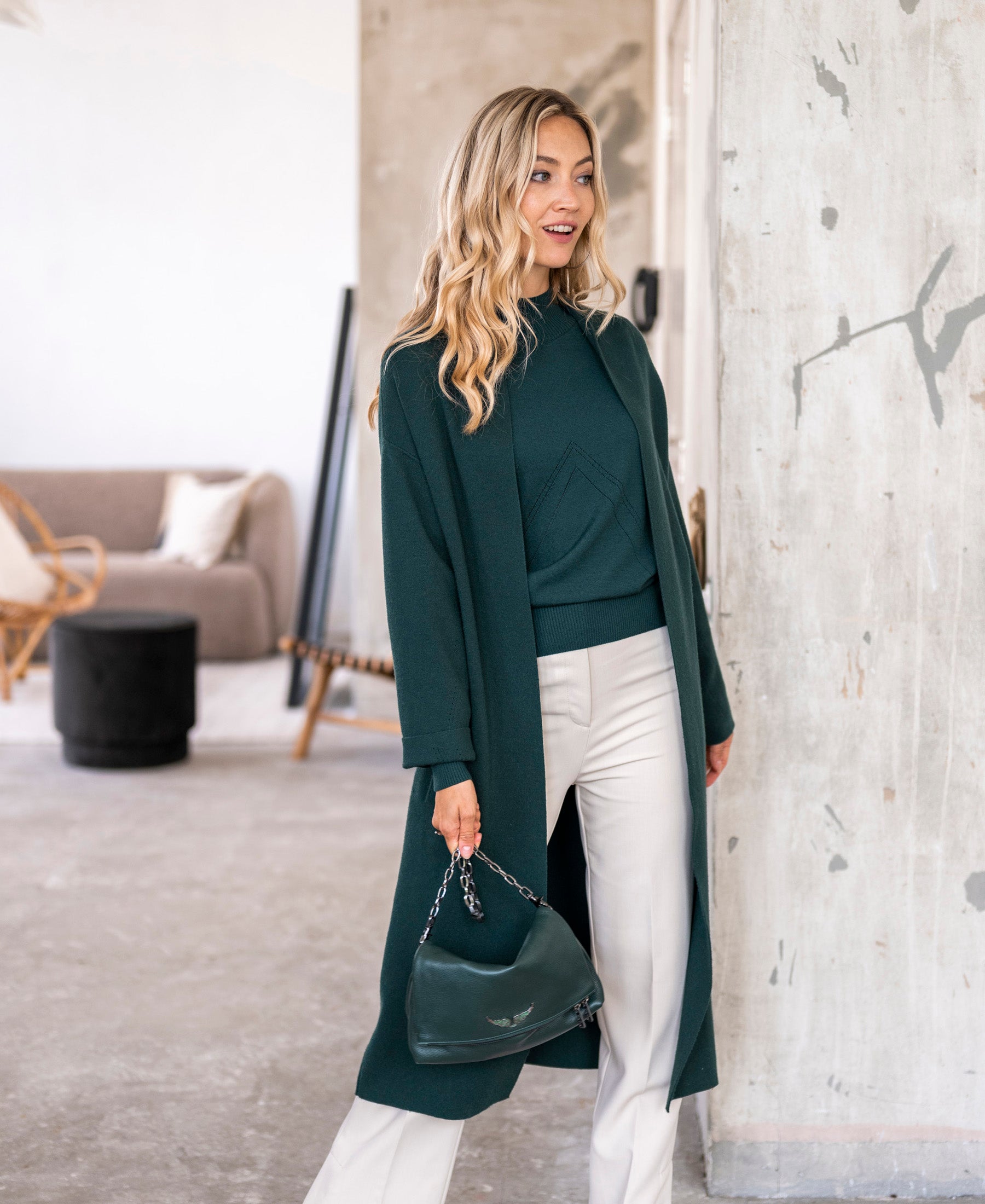 Dark green turtleneck sweater by PLEIN PUBLIQUE, shown on a woman standing in front of a textured wall.