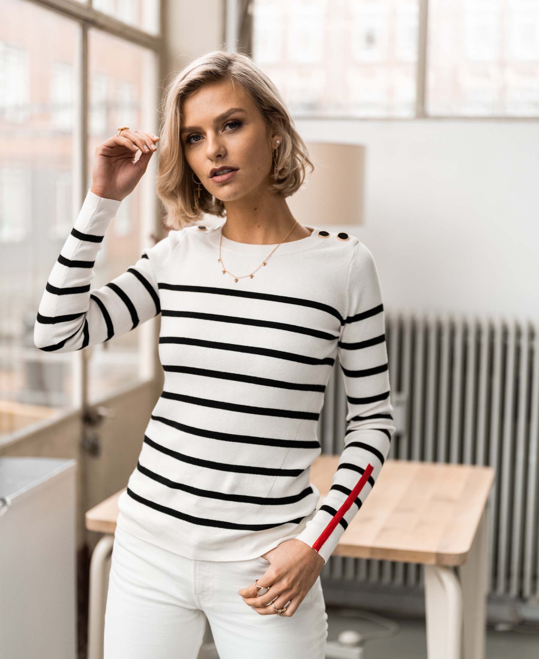 Woman wears the Breton striped sweater L'ELISA in ivory-black by PLEIN PUBLIQUE, posing near a table and looking at the camera.