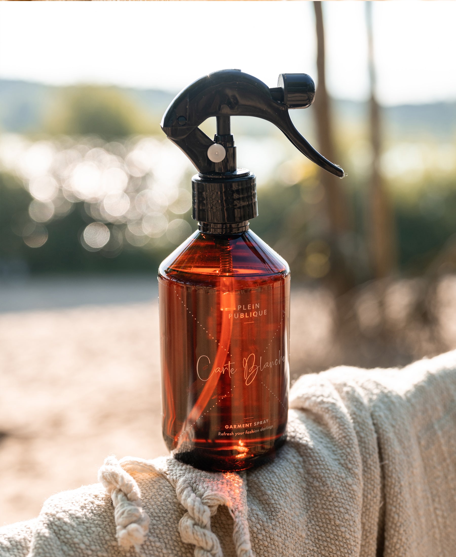 PLEIN PUBLIQUE Clothing fragrance spray bottle with black nozzle on beige fabric in sunlight.