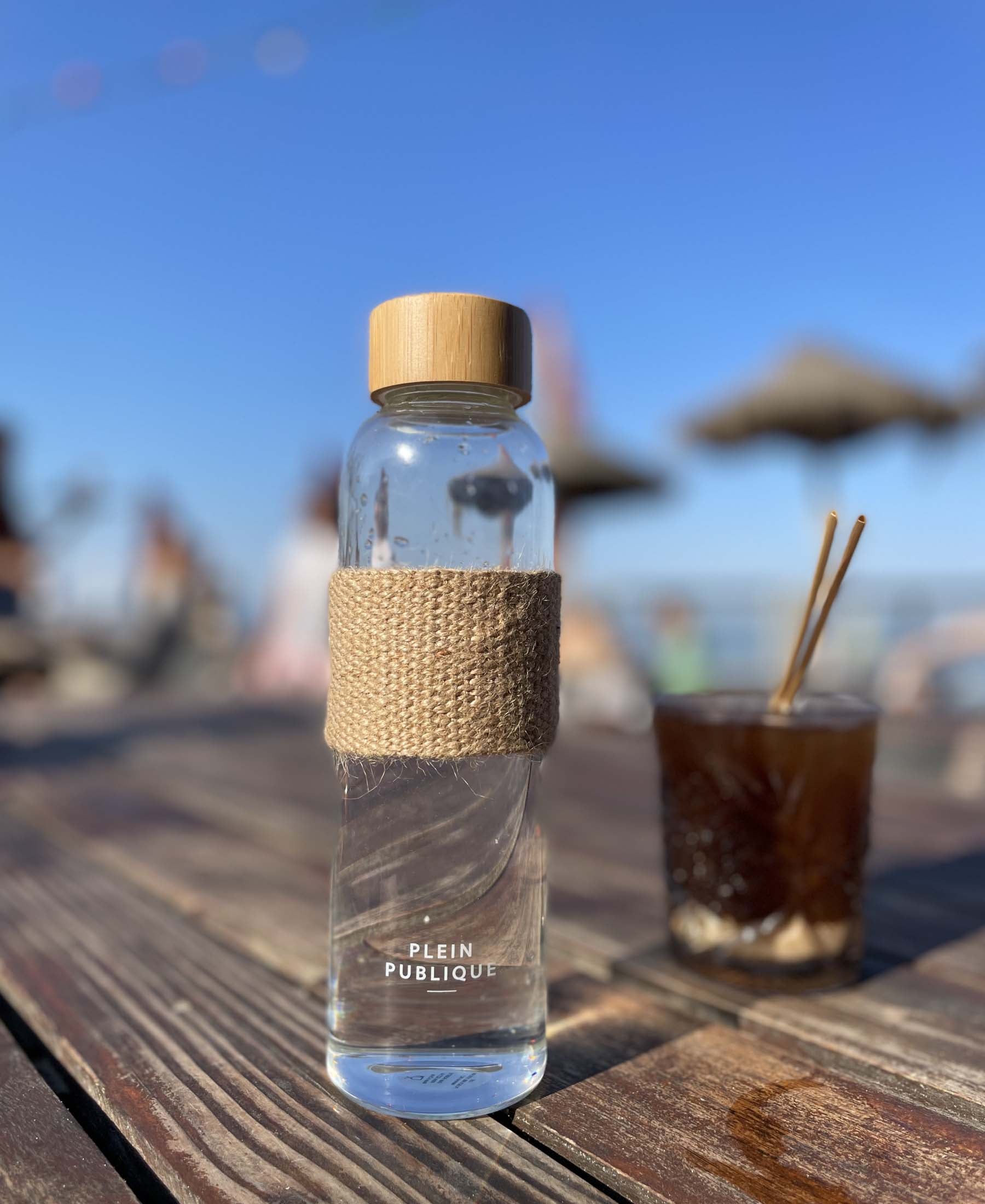 The glass water bottle with bamboo cap by PLEIN PUBLIQUE lies on a wooden table.