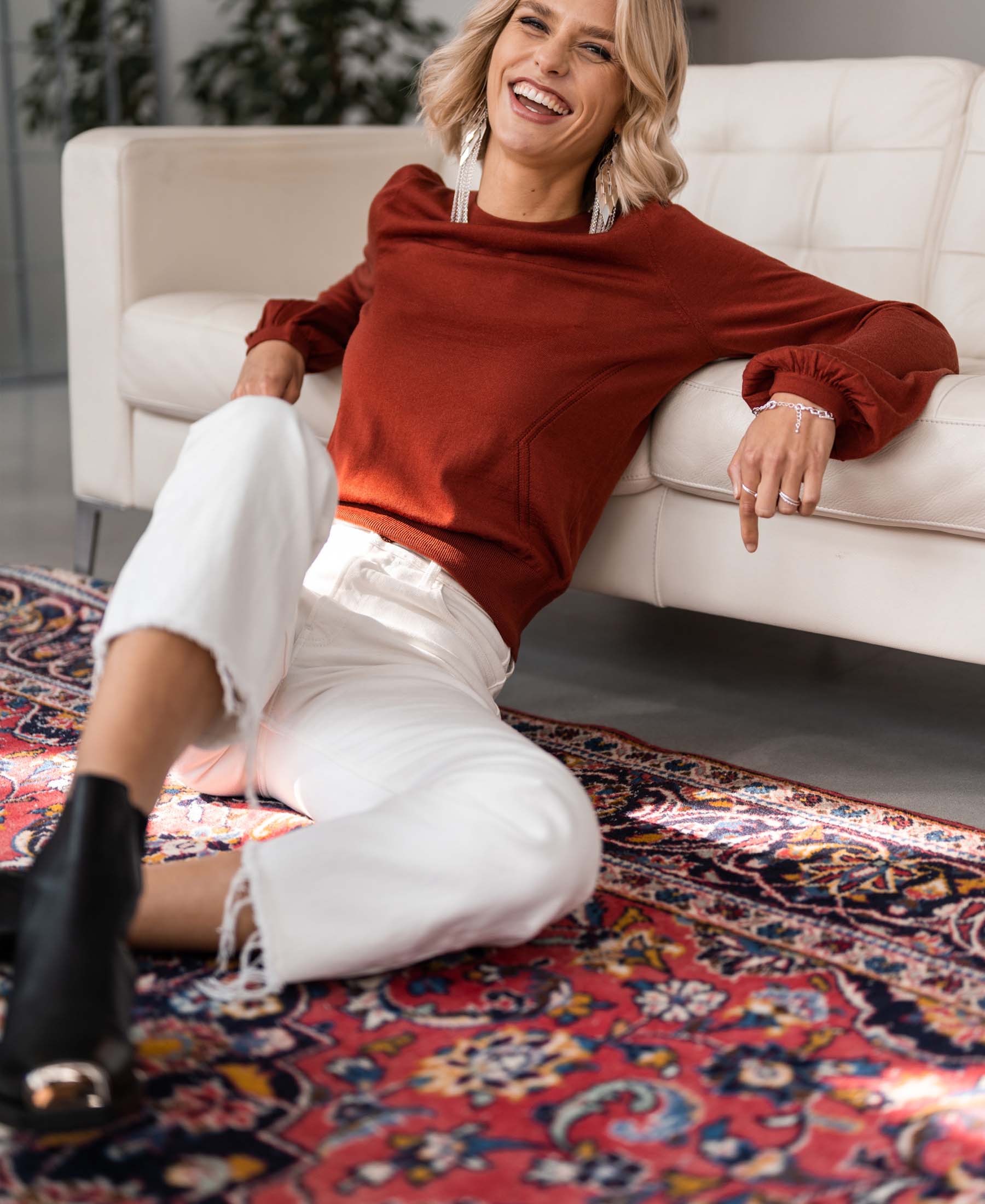Woman wearing the LA COEUR Rosewood Merino sweater by PLEIN PUBLIQUE, sitting on a rug near the sofa.