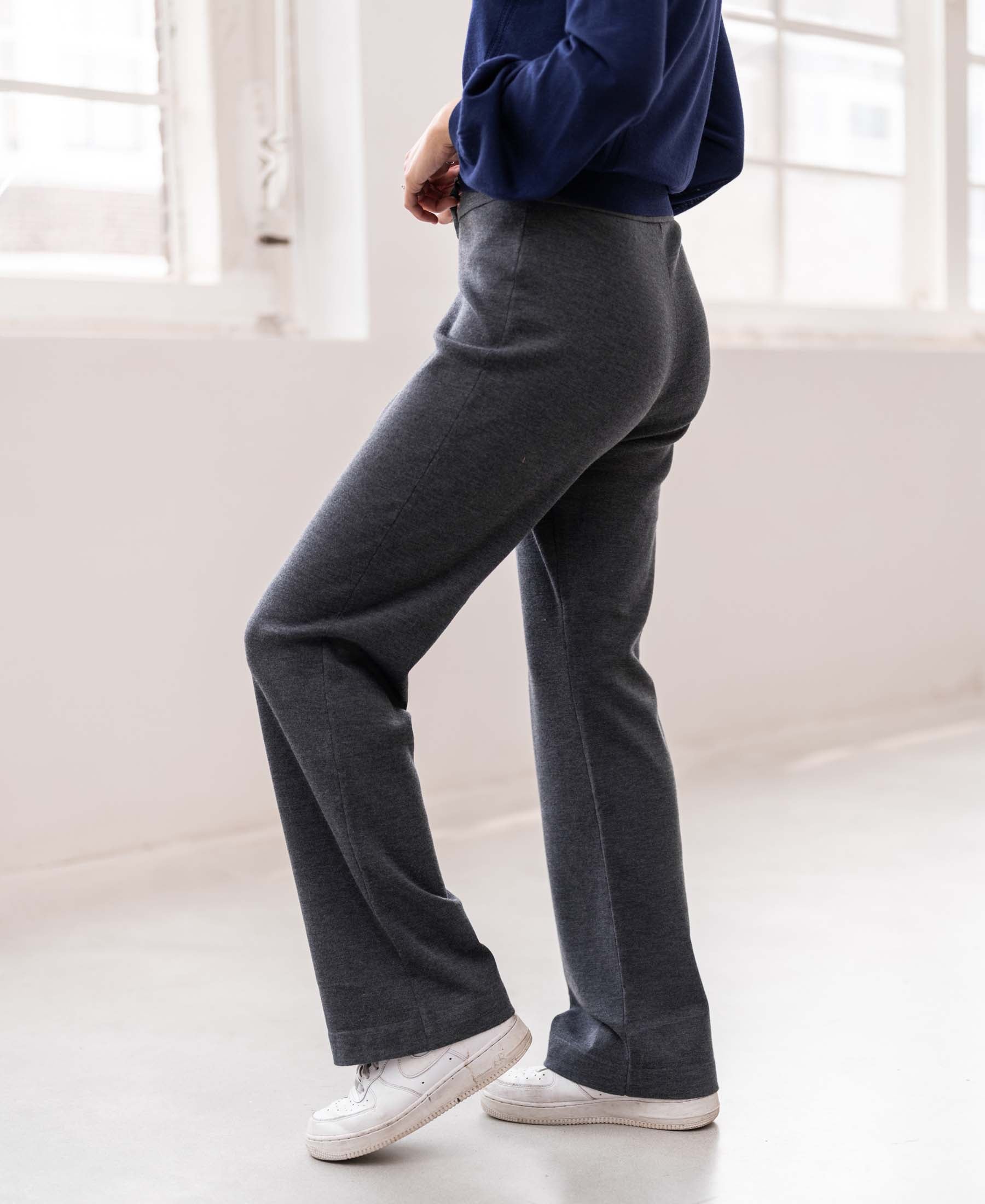 Woman wearing PLEIN PUBLIQUE knitted trousers LE LOURDES in anthracite stands by large windows indoors.