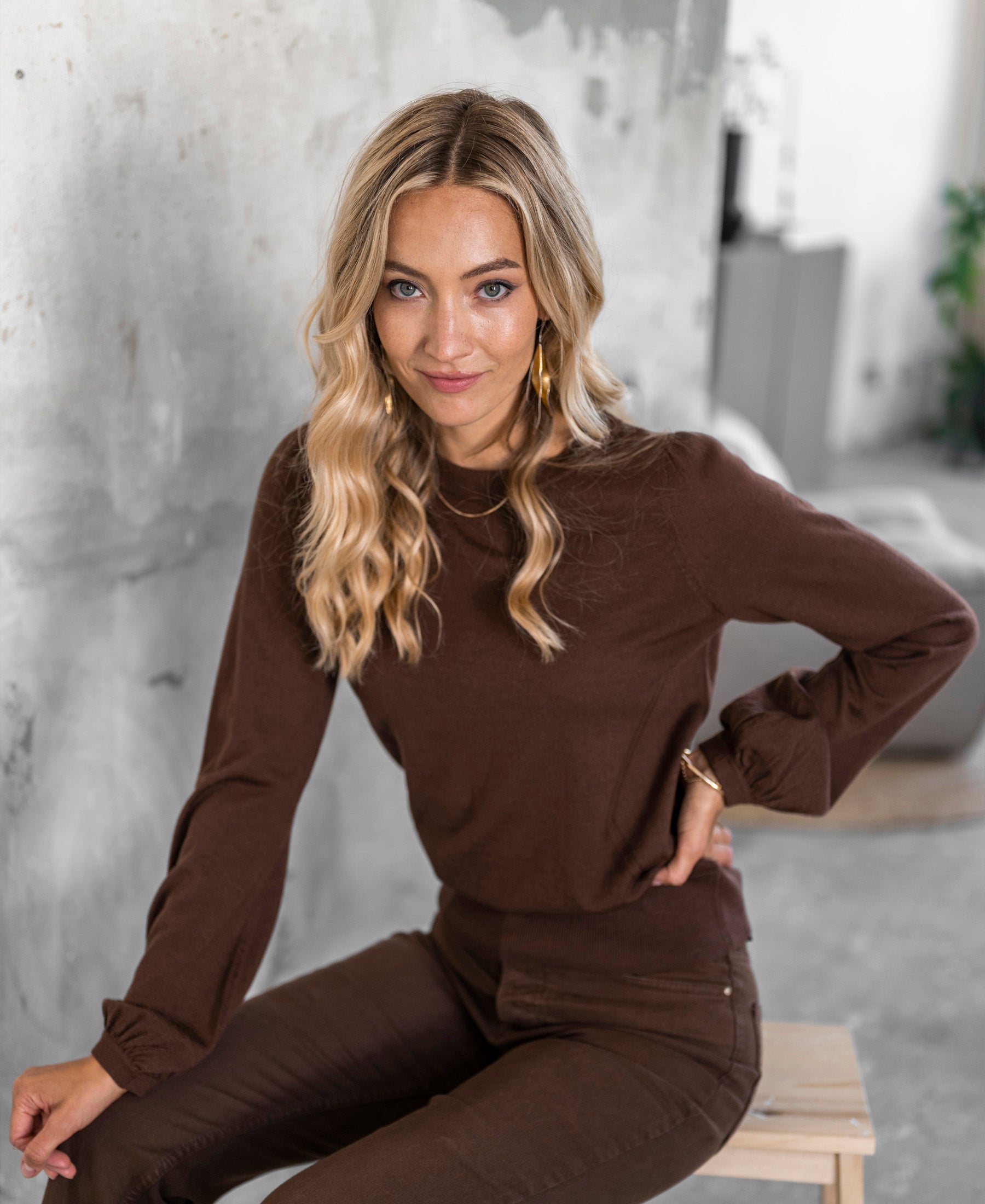 Woman wearing the brown LA COEUR Merino sweater by PLEIN PUBLIQUE, shown in a modern interior.