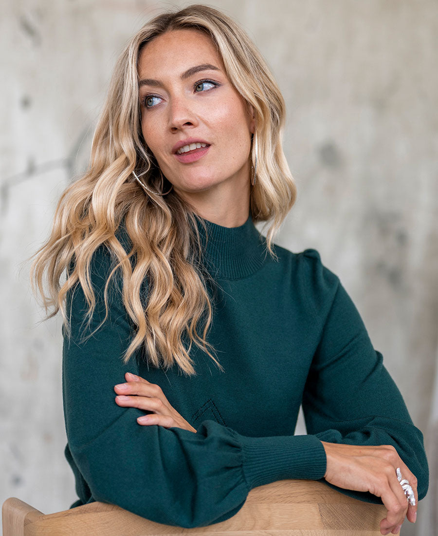 A woman wears a dark green turtleneck sweater, seated and smiling slightly.