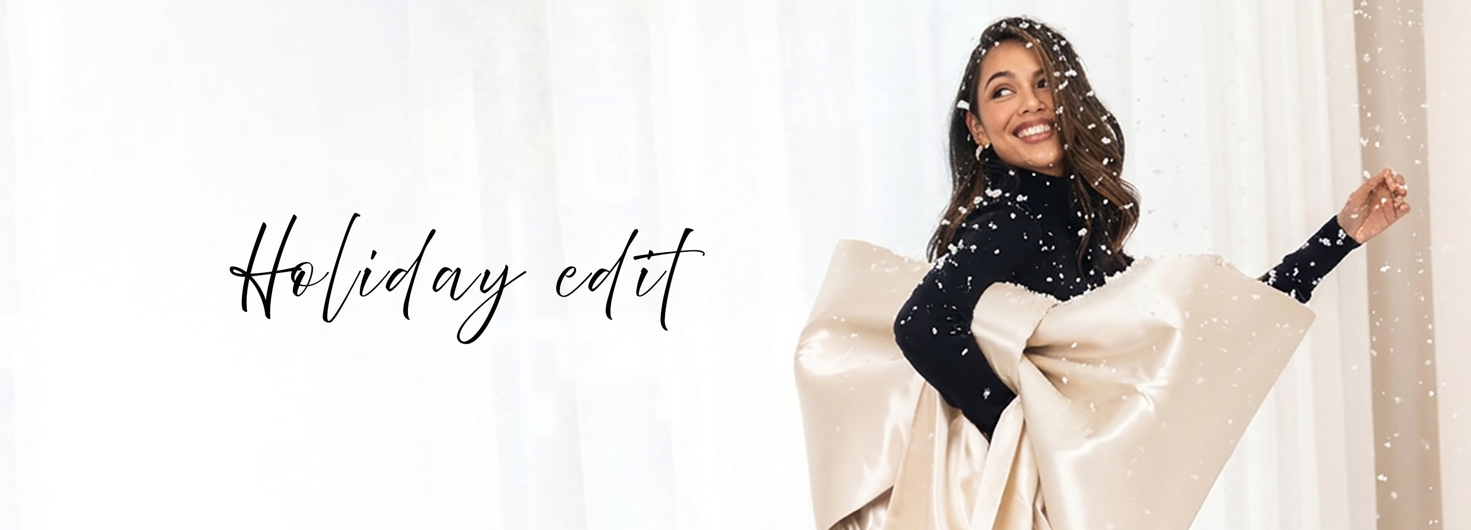 Large cream-colored fabric bow held up close, with falling snowflakes and the words Holiday edit in elegant script. White curtains are visible in the background.