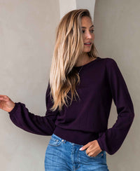Woman wearing a red Merino wool sweater LA COEUR by PLEIN PUBLIQUE stands by a light wall, looking to the side.