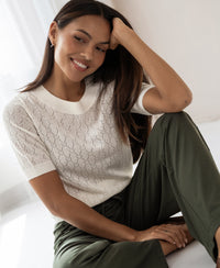Woman wearing ivory sweater with heart pattern, arm on knee. Sweater by PLEIN PUBLIQUE, style LE BATTANT.