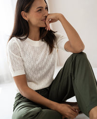 Woman wearing an ivory sweater with heart pattern from PLEIN PUBLIQUE LE BATTANT sits smiling on the floor in a bright room.