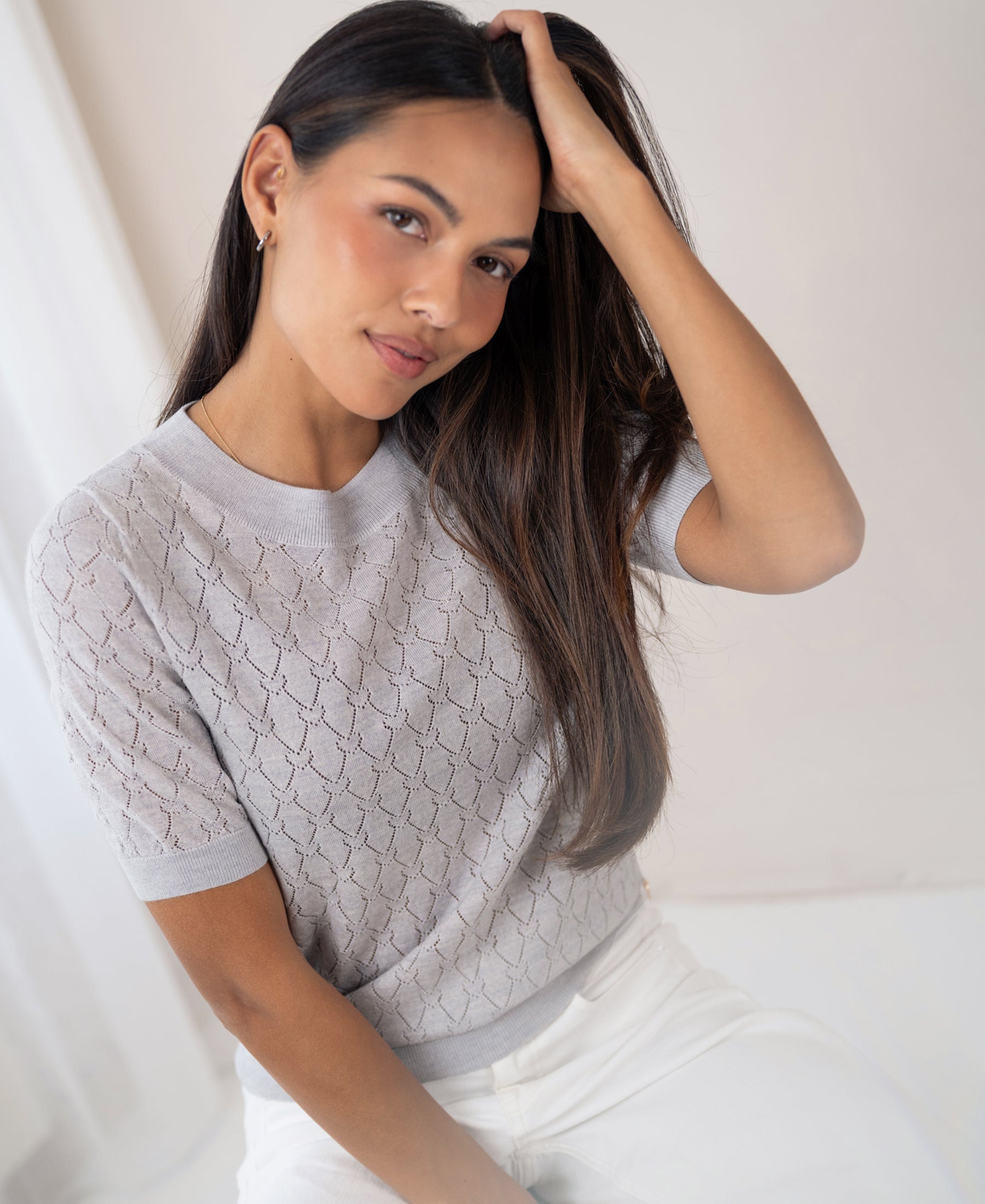 Light grey sweater with heart pattern, shown on a woman with long dark hair, sitting and smiling slightly.