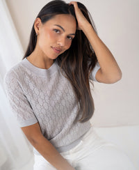 Light grey sweater with heart pattern, shown on a woman with long dark hair, sitting and smiling slightly.