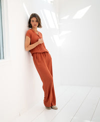 Terra-colored LA LYNETTE pants by OVERIG shown on a model leaning against a white wall in a sunlit, minimalist room.