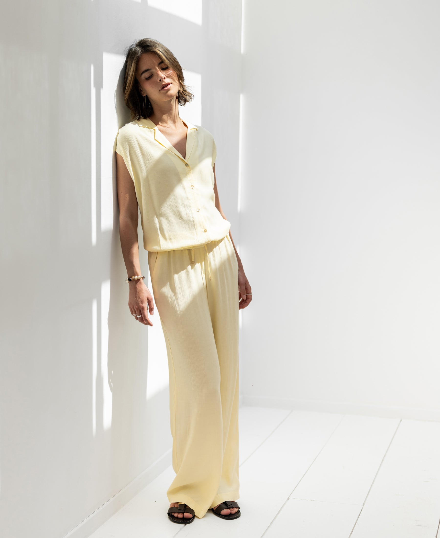 Sleeveless blouse LA BIJOU soft yellow by OVERIG, shown worn, model leans against sunlit wall with eyes closed.