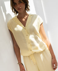 Sleeveless blouse in soft yellow by OVERIG, worn by a woman standing in sunlight against a white wall.