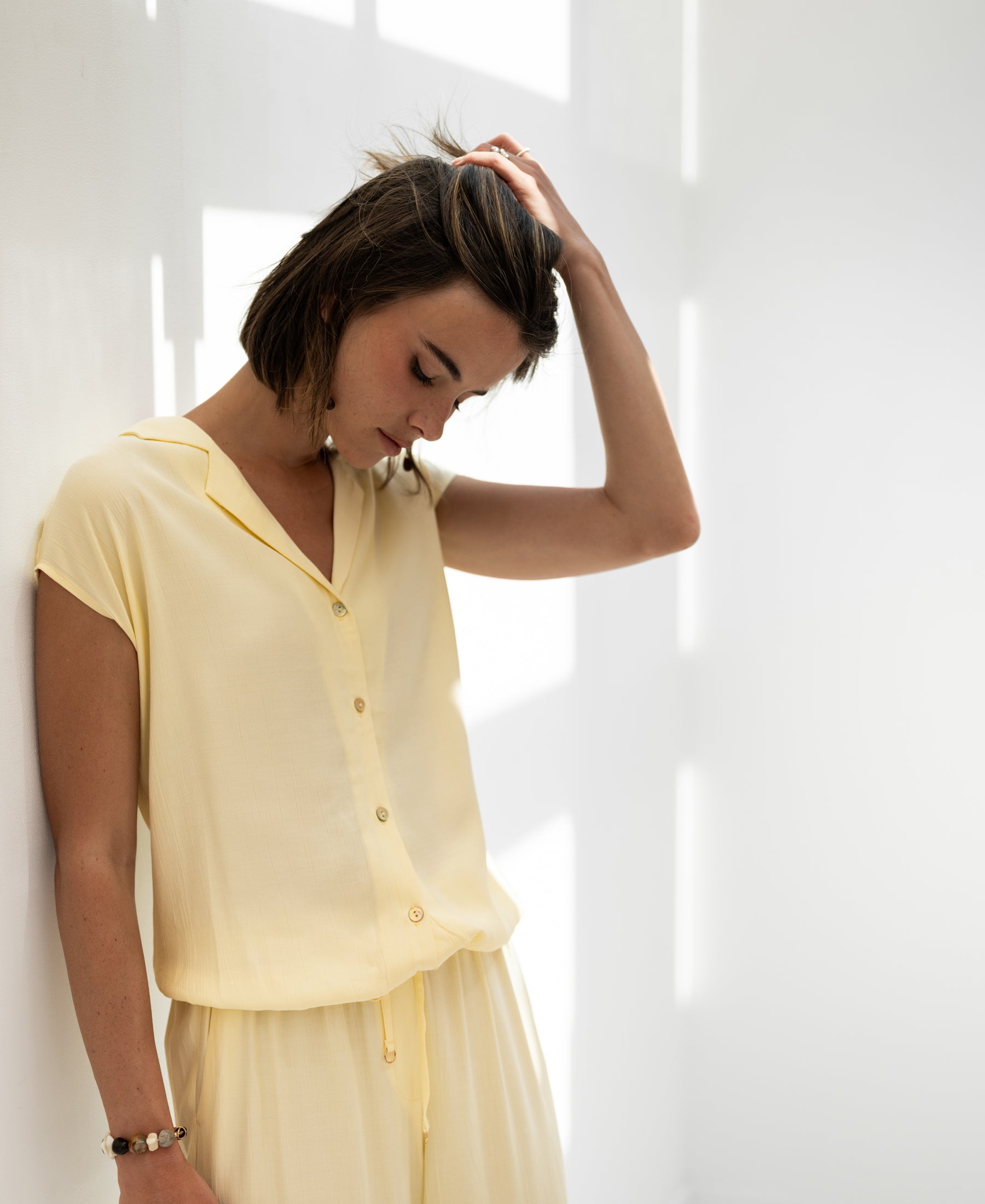 Sleeveless soft yellow La Bijou blouse shown on woman standing by a sunlit wall.