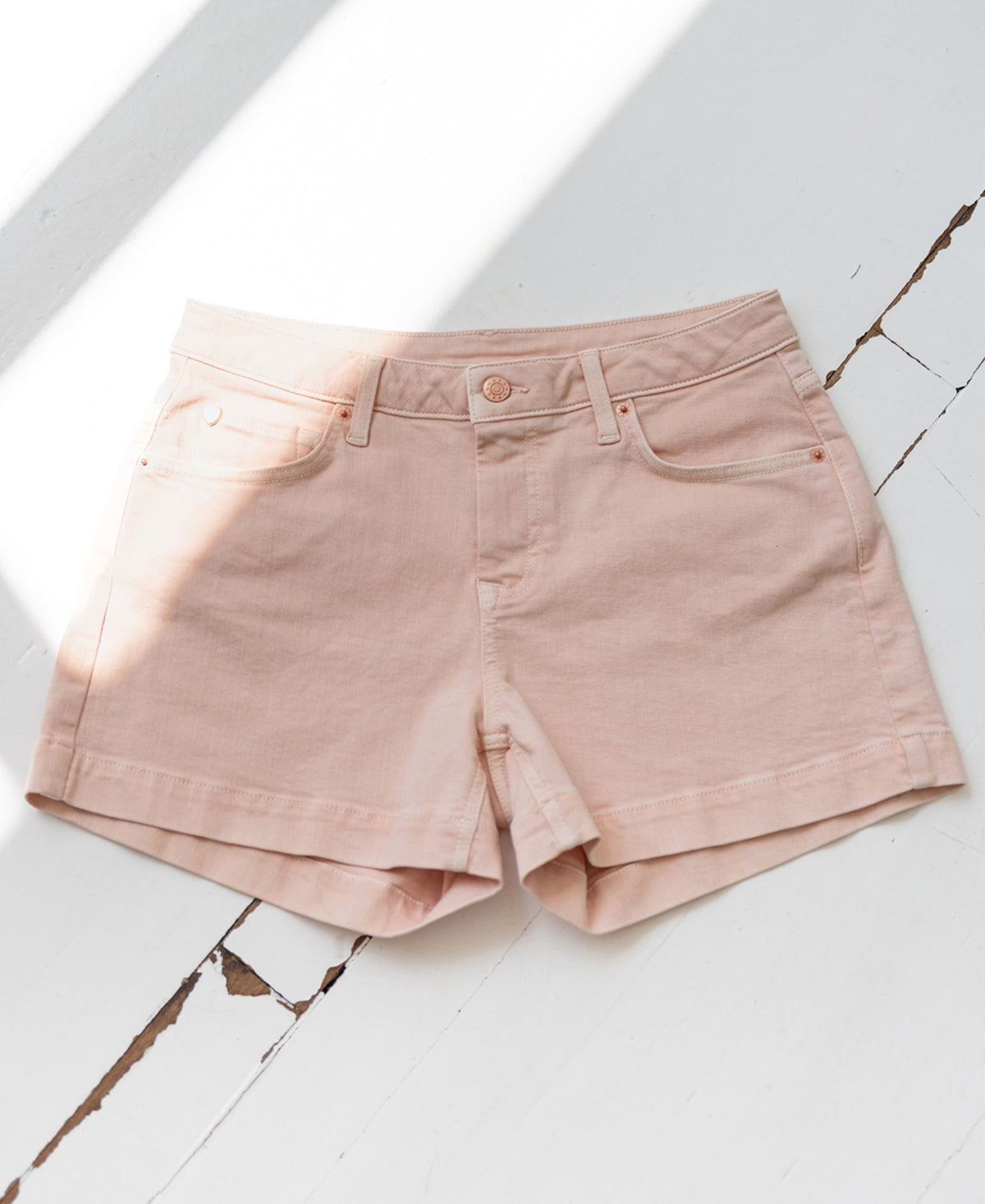 Blush 5-pocket shorts lie flat on a sunlit, cracked white floor with soft shadows.