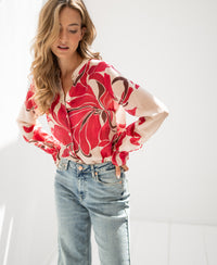 Woman wearing a red print PLEIN PUBLIQUE blouse called The Binocle, standing in a sunlit room.