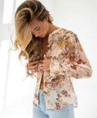 Woman wearing Plein Publique blush floral print blouse stands in natural light, looking down.