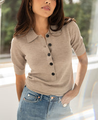Woman wearing a dark sand-colored merino wool polo shirt by PLEIN PUBLIQUE stands indoors with one hand in her pocket.