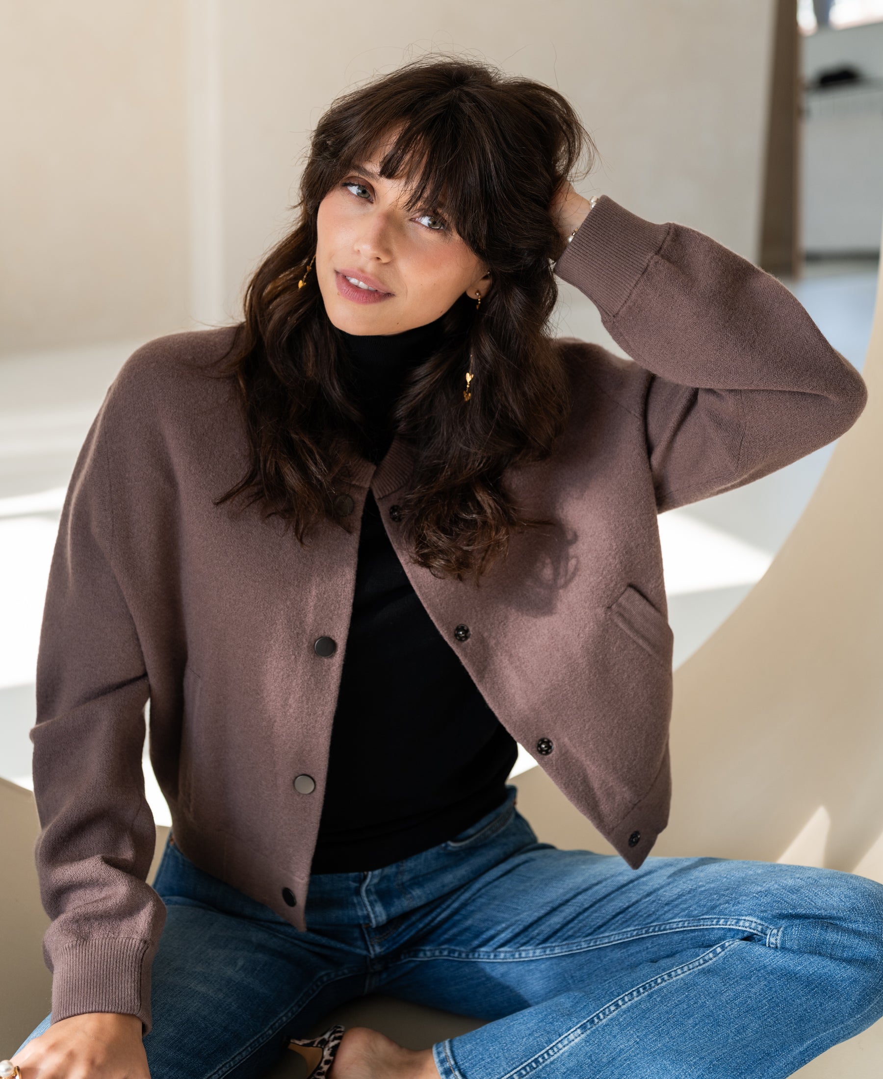 Woman wearing the PLEIN PUBLIQUE LA BOMBA bomber jacket in Innuendo, sitting indoors and smiling softly.
